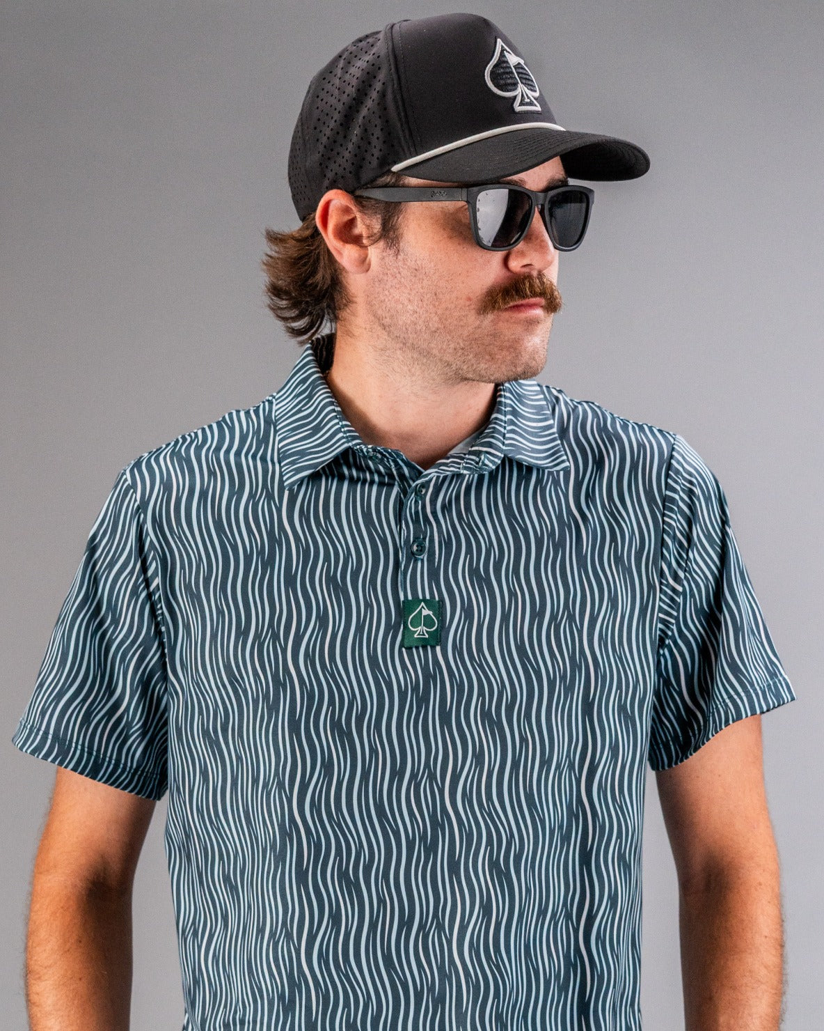 Male model wearing a black cap with a spade logo, black sunglasses, and a blue and white wavy patterned golf shirt.