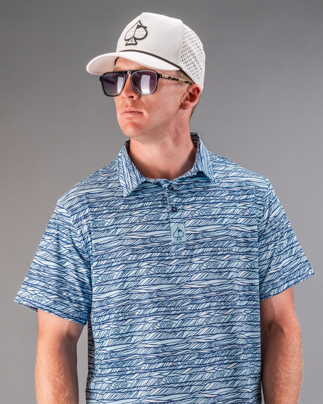 Man wearing a blue and white patterned golf shirt, white cap with spade logo, and dark sunglasses, showcasing bold golf st...