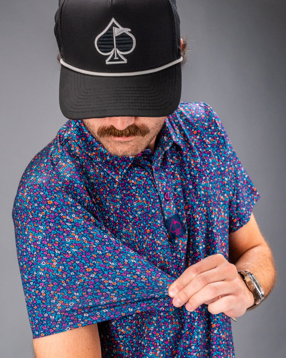 Lavender Flower Power golf polo featuring a vibrant floral pattern, worn by a man adjusting the sleeve.