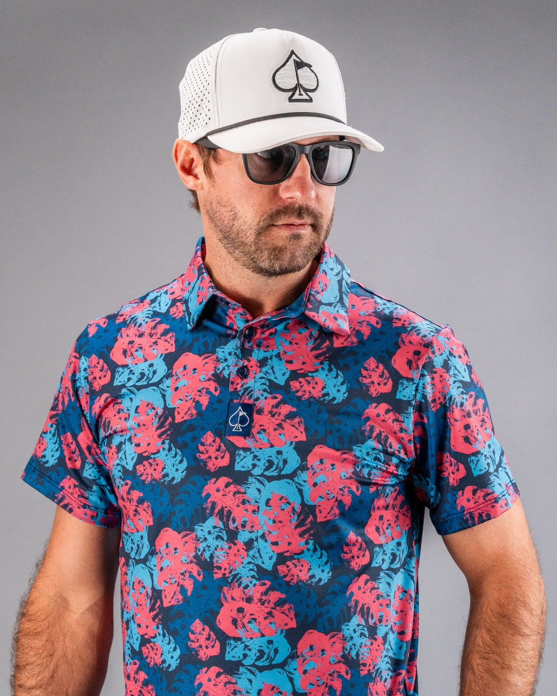 Man wearing a white mesh baseball cap with a black spade logo, black sunglasses, and a colorful tropical golf shirt with p...