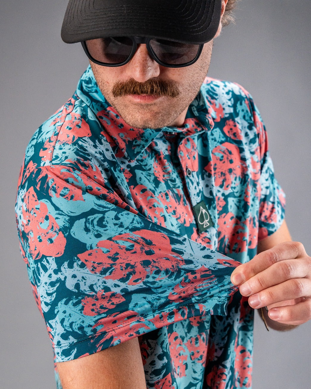 Man wearing a bold, colorful floral golf shirt in blue and coral, with a black cap and sunglasses, showcasing unique golf ...