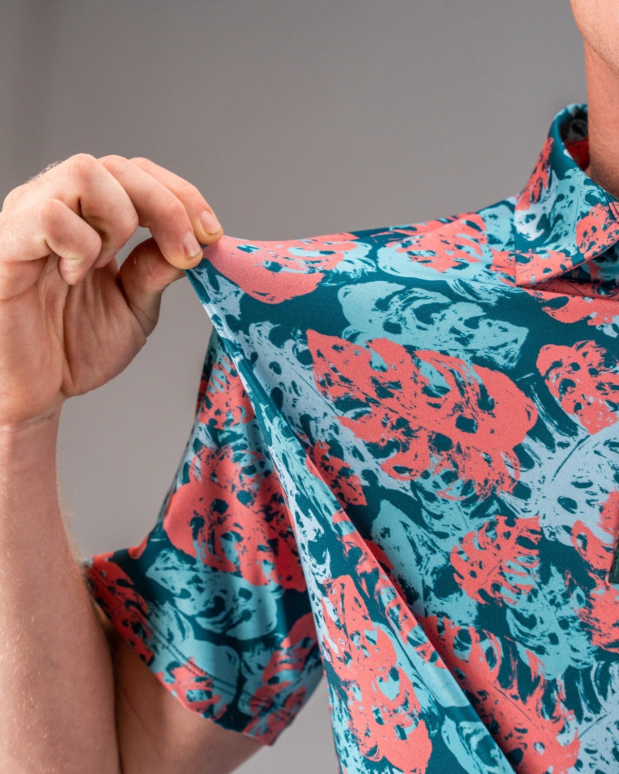 Bold teal shirt featuring a vibrant painted monstera leaf pattern in coral and blue, perfect for stylish golfers.