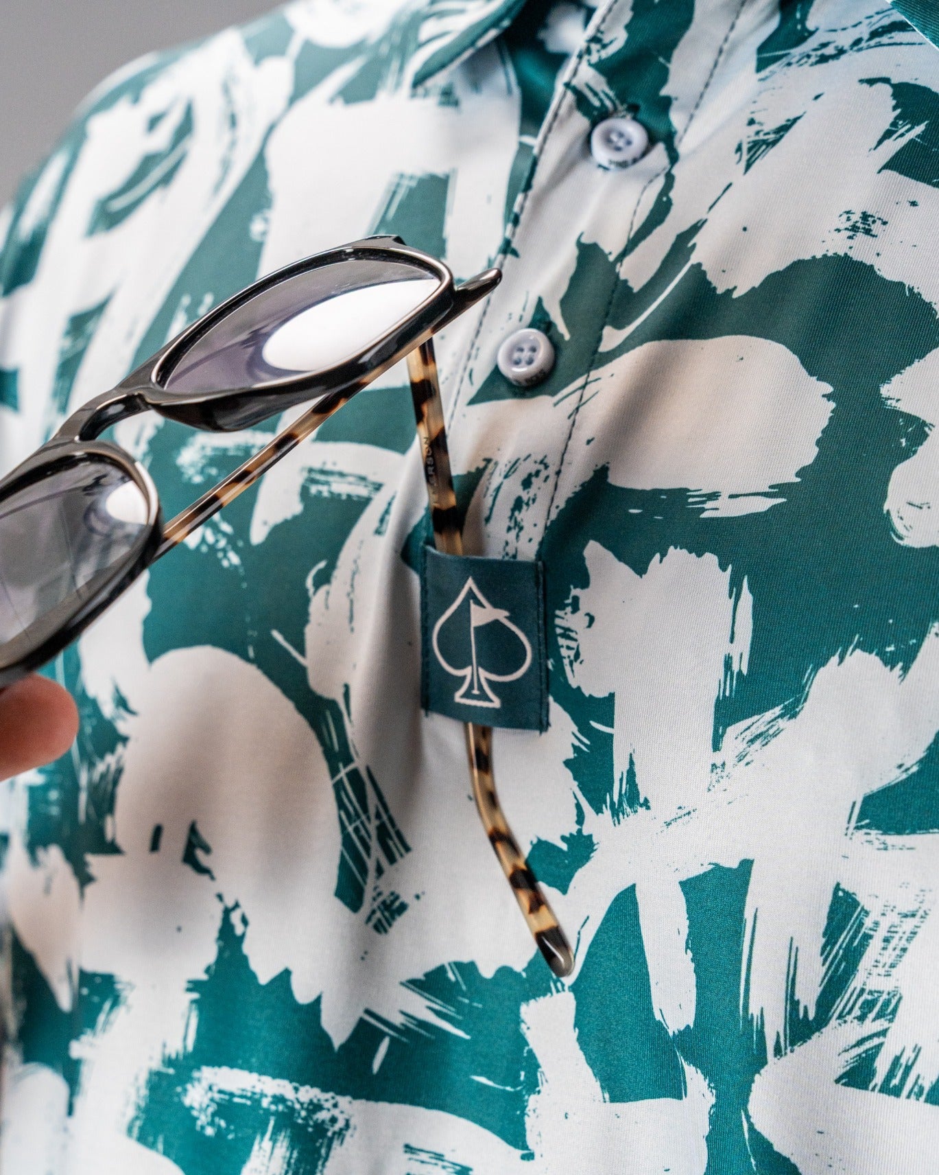 Close-up of a green and white patterned polo shirt with a spade logo, featuring sunglasses held in hand.