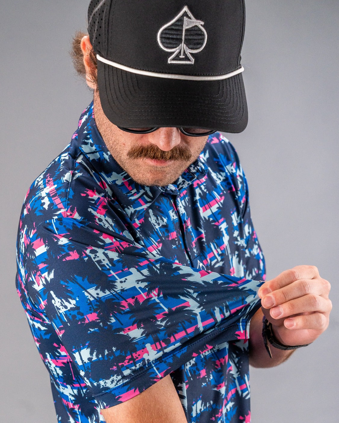 Man wearing a bold, colorful golf shirt with palm tree pattern, black cap with spade logo, adjusting his sleeve.
