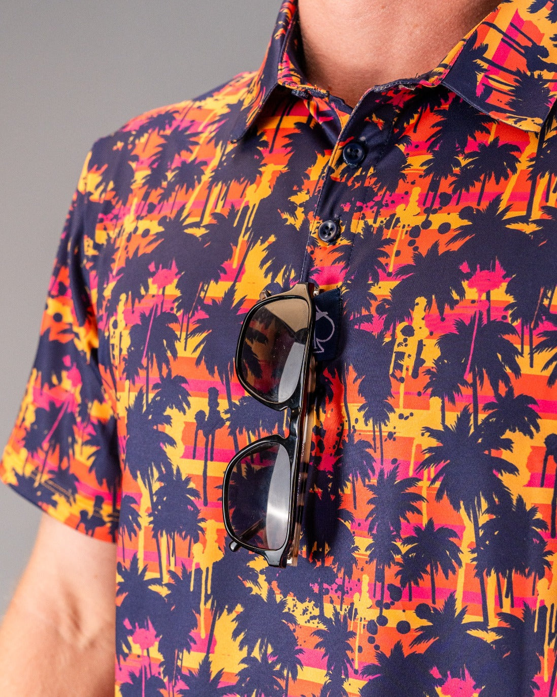Close-up of a person wearing a vibrant, tropical palm tree patterned golf shirt in navy, pink, orange, and yellow. Sunglas...