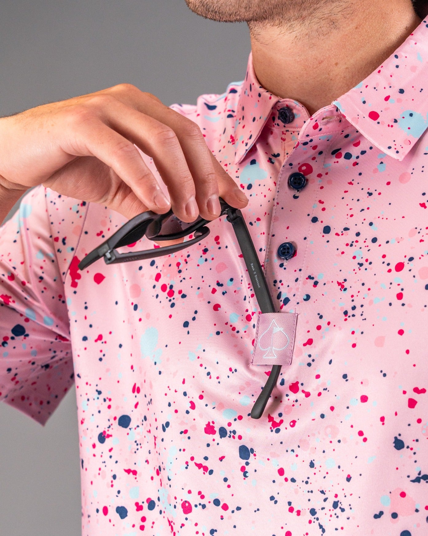 Men's pink splatter golf polo featuring vibrant paint splashes, paired with sunglasses held at the collar.