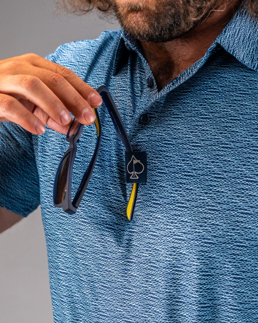 Men's blue golf polo shirt with a textured squiggle pattern, featuring a logo tag and sunglasses held in hand.