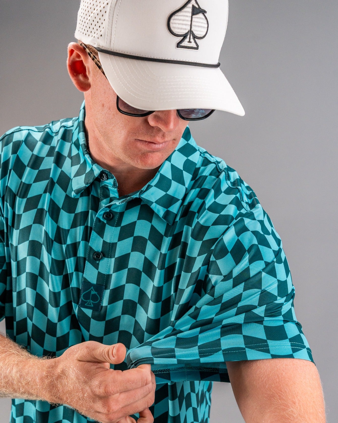 Bold wavy checkered golf shirt in teal, featuring a logo on the chest, worn by a golfer adjusting the sleeve.