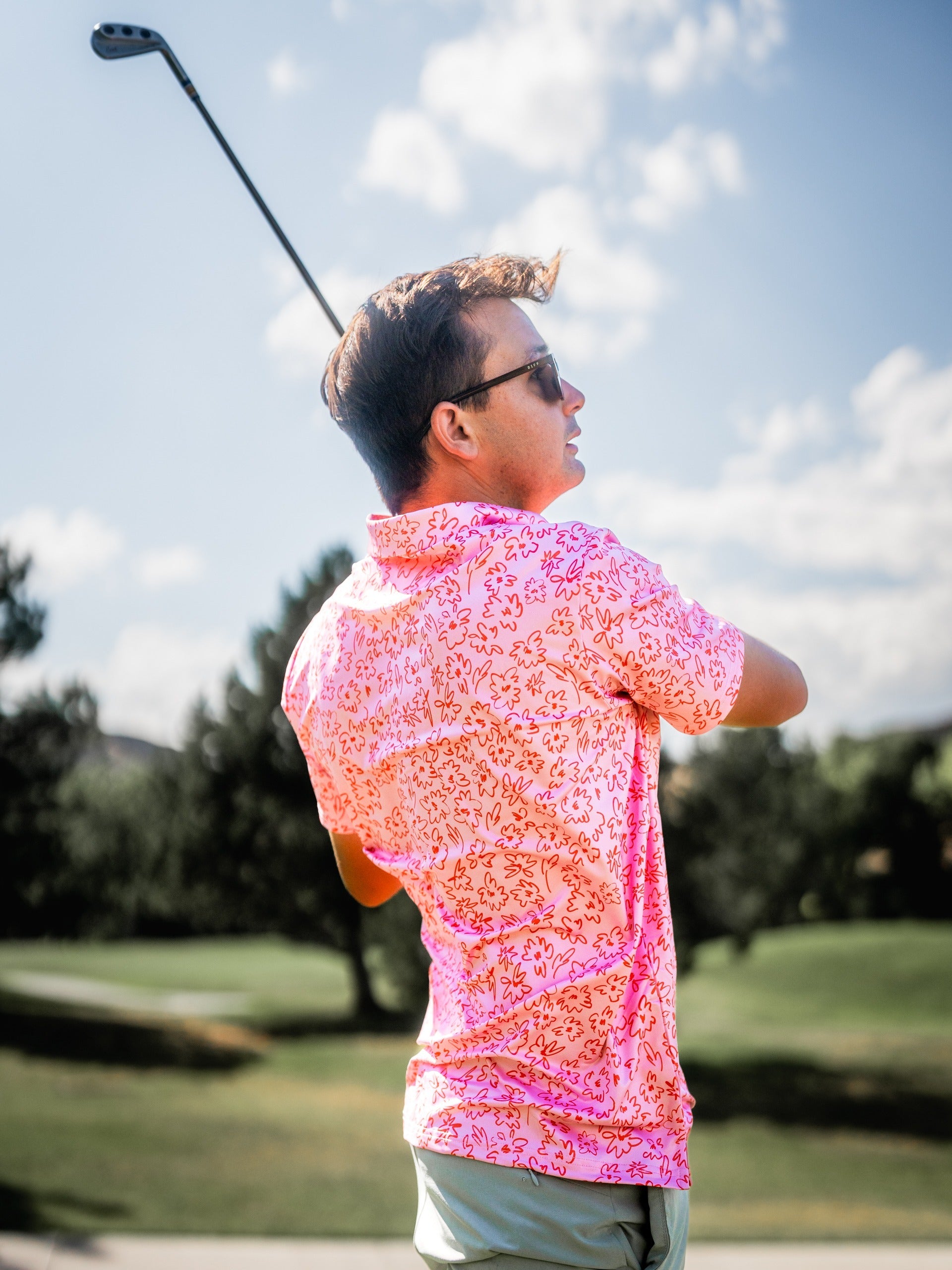 A golfer swings a club while wearing a vibrant pink shirt with a floral doodle pattern, set against a sunny golf course backdrop.