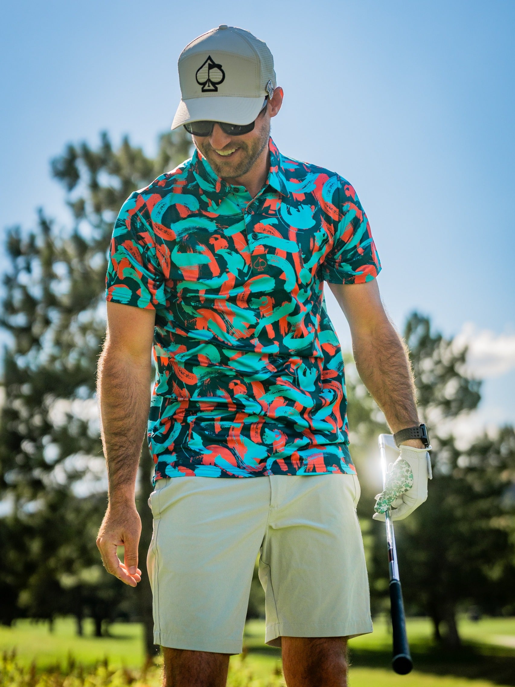 Man wearing a vibrant Paint Strokes Coral golf shirt with colorful patterns, standing on a golf course, holding a club.