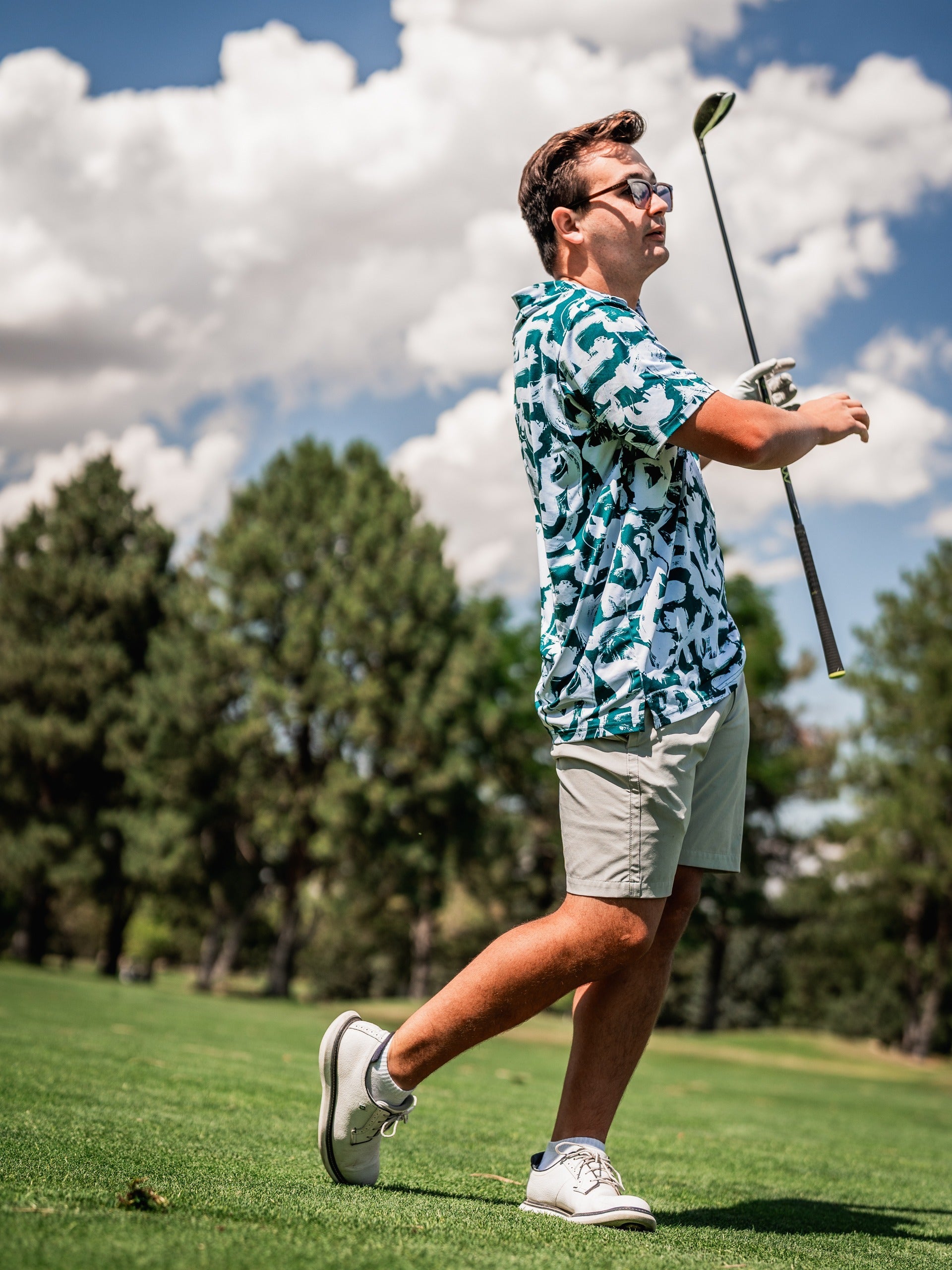 A golfer in a vibrant green paint strokes patterned shirt swings a club on a sunny course, showcasing bold golf style.