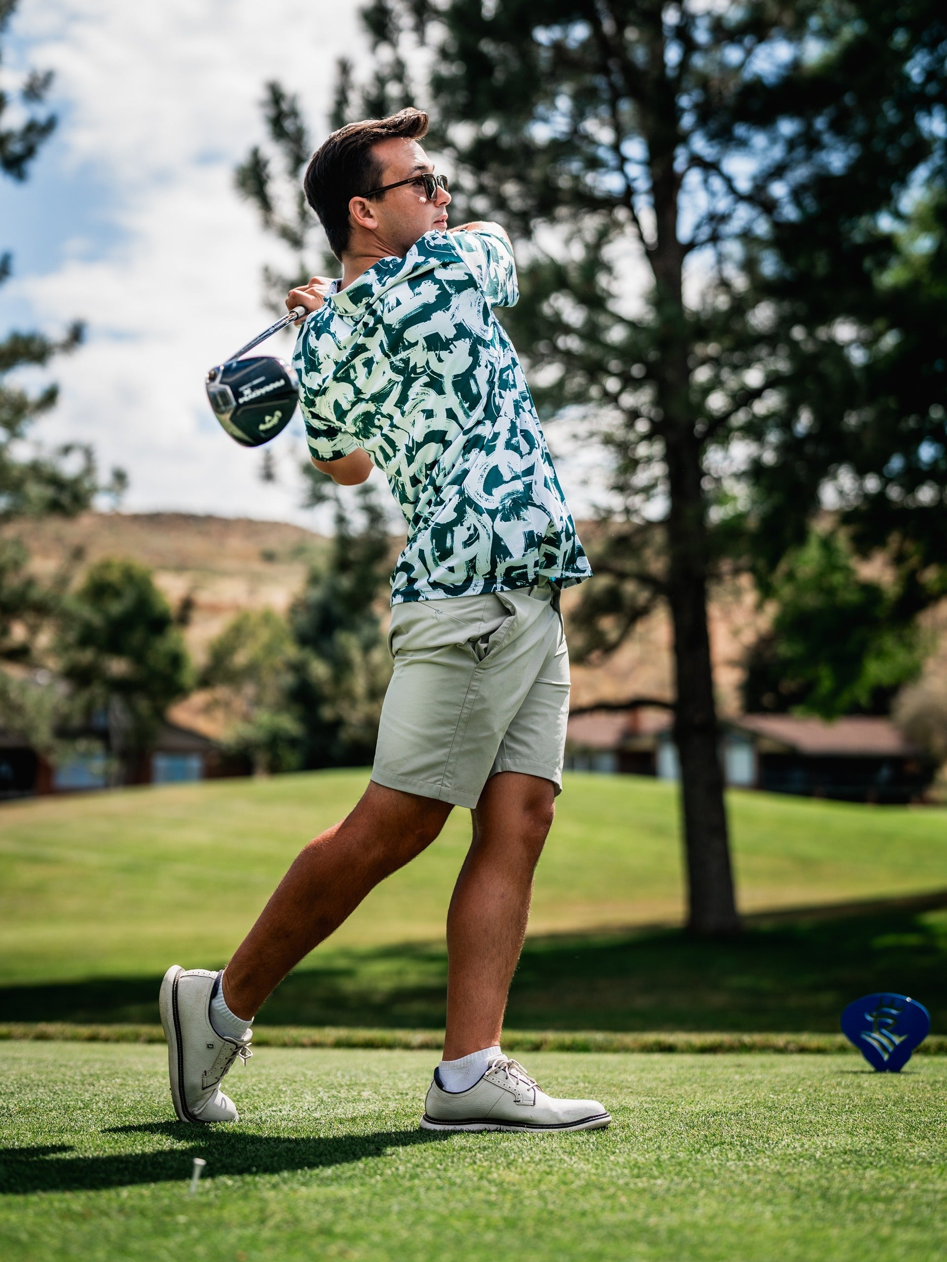 A golfer swings a club on the course, wearing a vibrant green and white patterned shirt and light shorts.