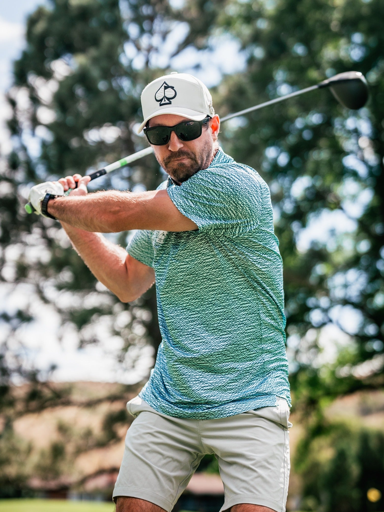 A golfer in a teal squiggle-patterned shirt and cap prepares to swing a golf club on a sunny course.