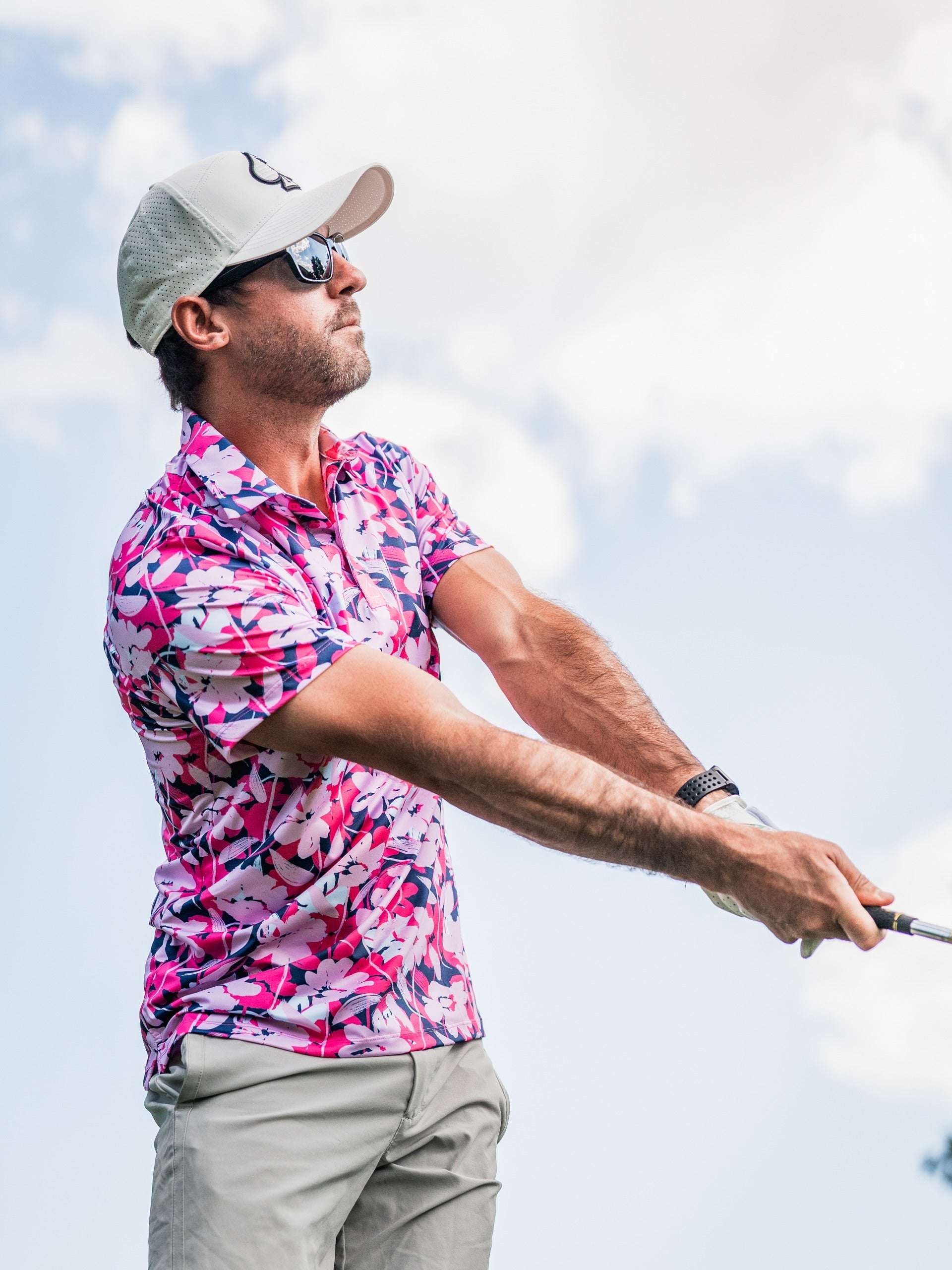 Man in pink floral golf shirt, beige cap and sunglasses swings golf club outdoors against cloudy sky.