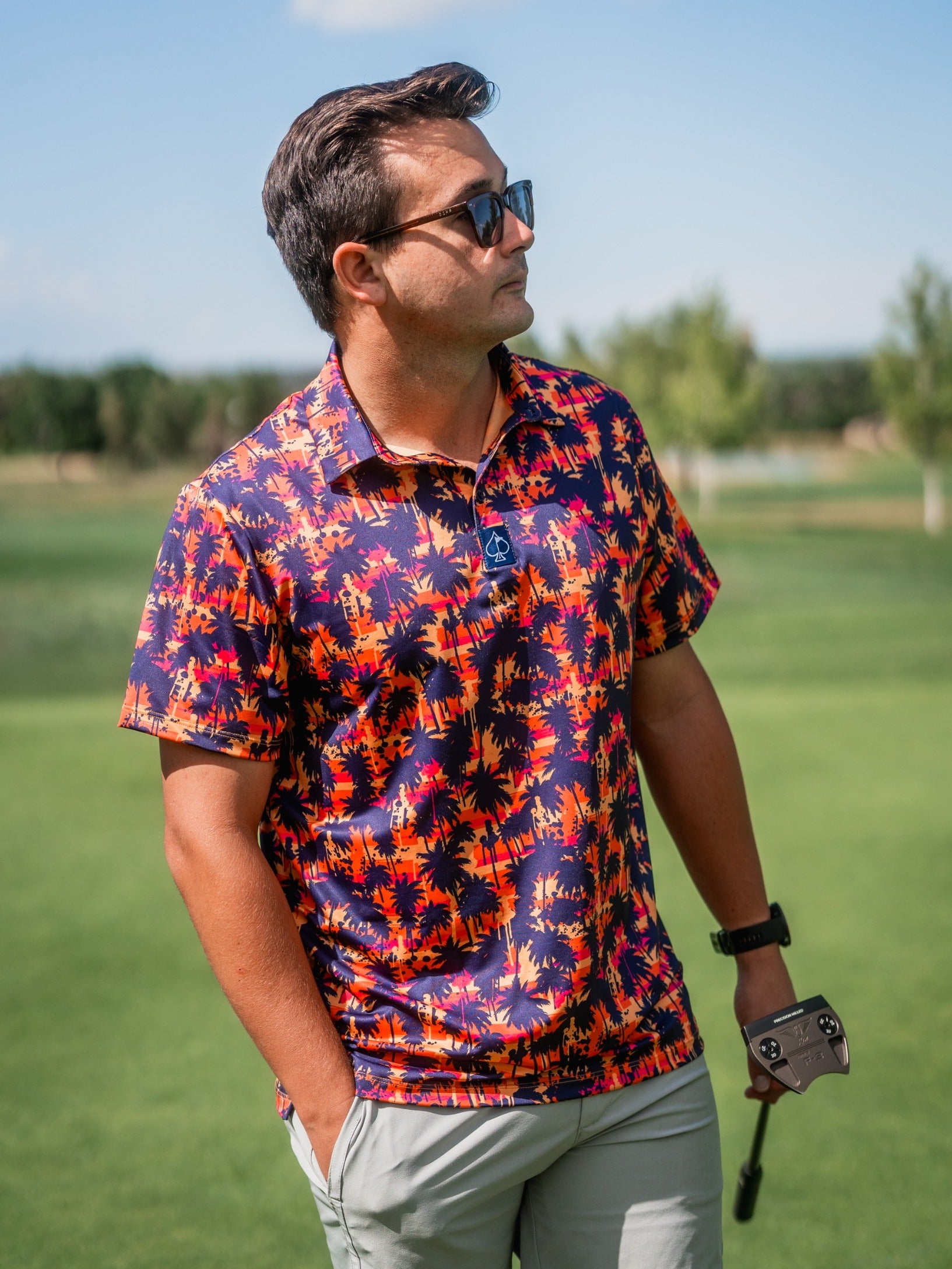 Man wearing a vibrant, tropical-themed golf shirt with palm trees in orange, navy, and pink, on a golf course.
