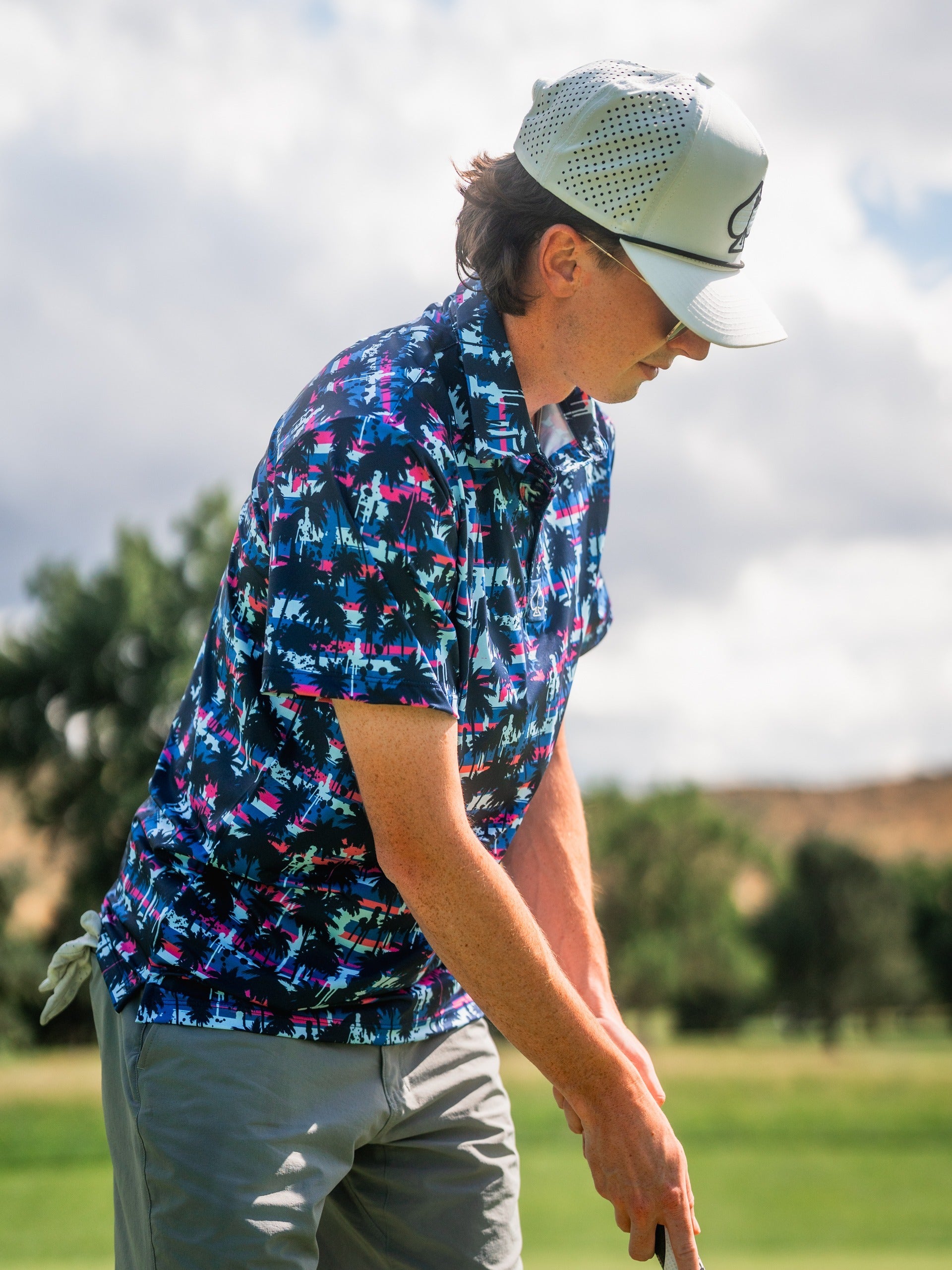 Male golfer in a vibrant blue, pink, and black tropical print shirt, beige pants, white cap, and sunglasses on a golf course.