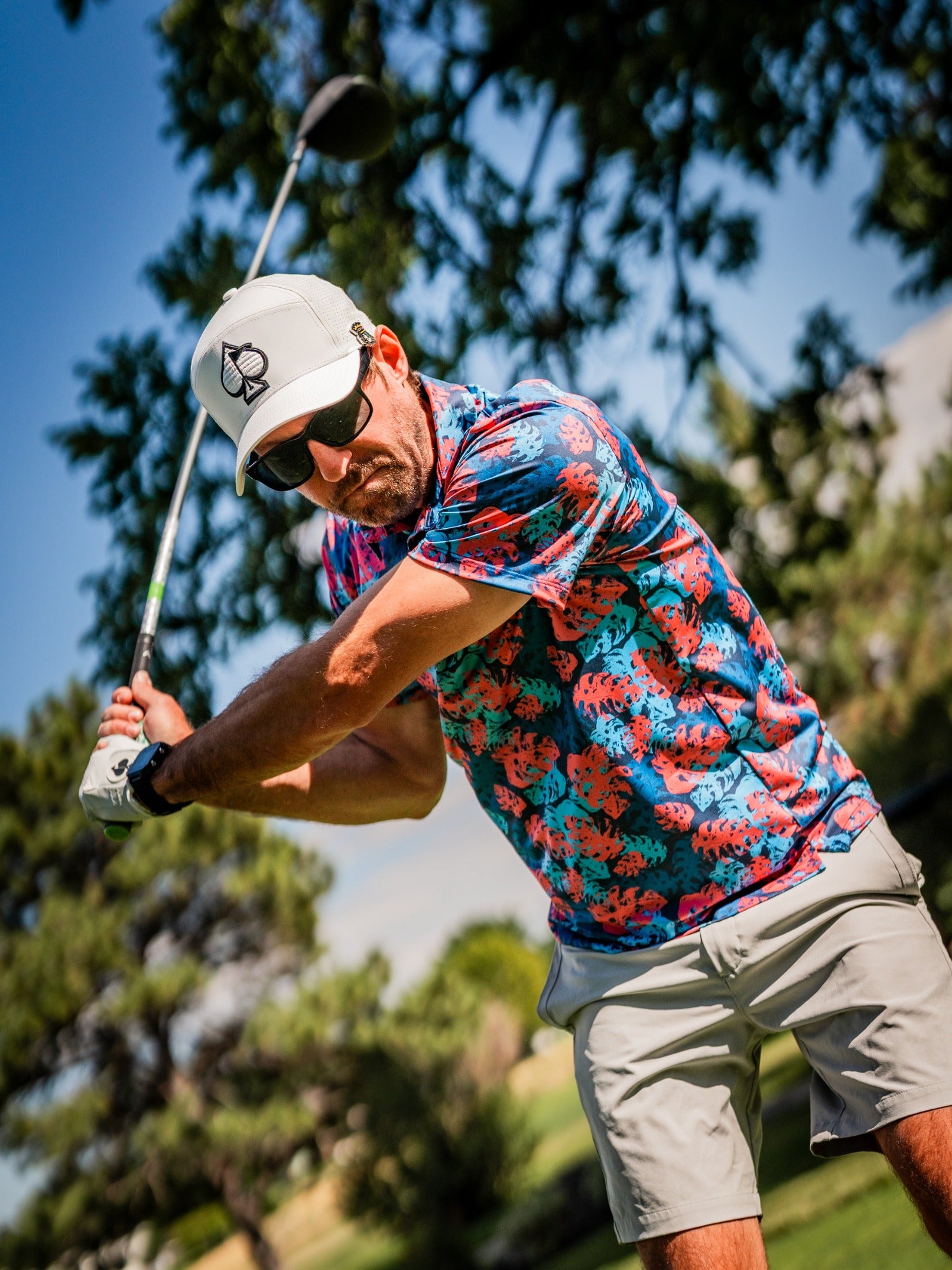 A golfer swings a club while wearing a vibrant blue and pink monstera-patterned shirt, showcasing bold style on the course.