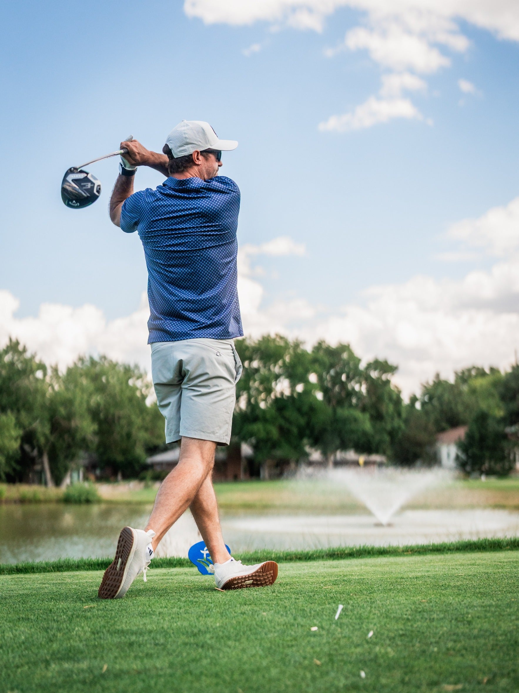 Male golfer in blue patterned polo, khaki shorts, white cap, swinging club on lush green golf course with water and trees.