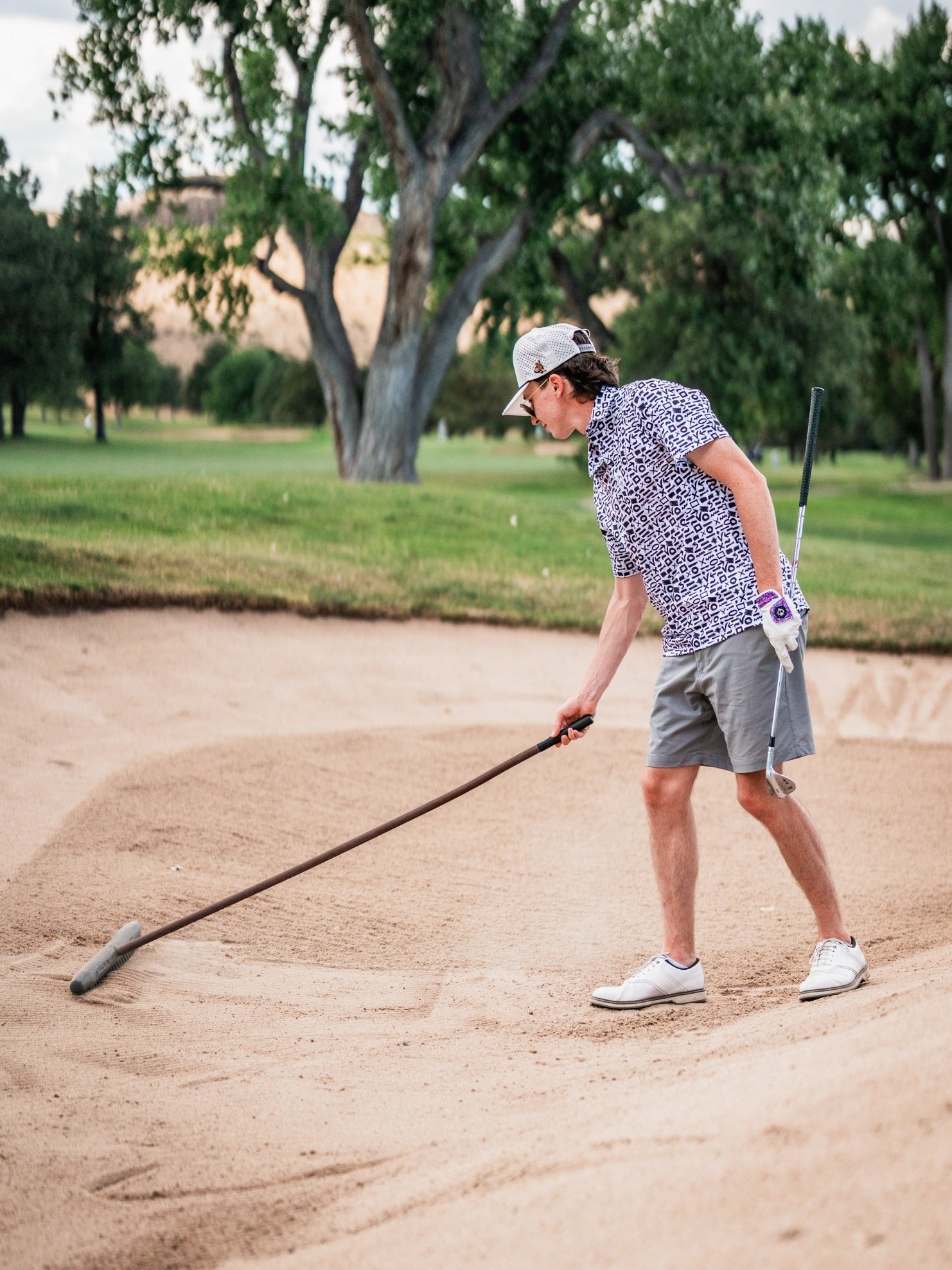 Golfer in a patterned shirt and cap cleaning a sand trap, with lush green trees in the background, on a sunny golf course.