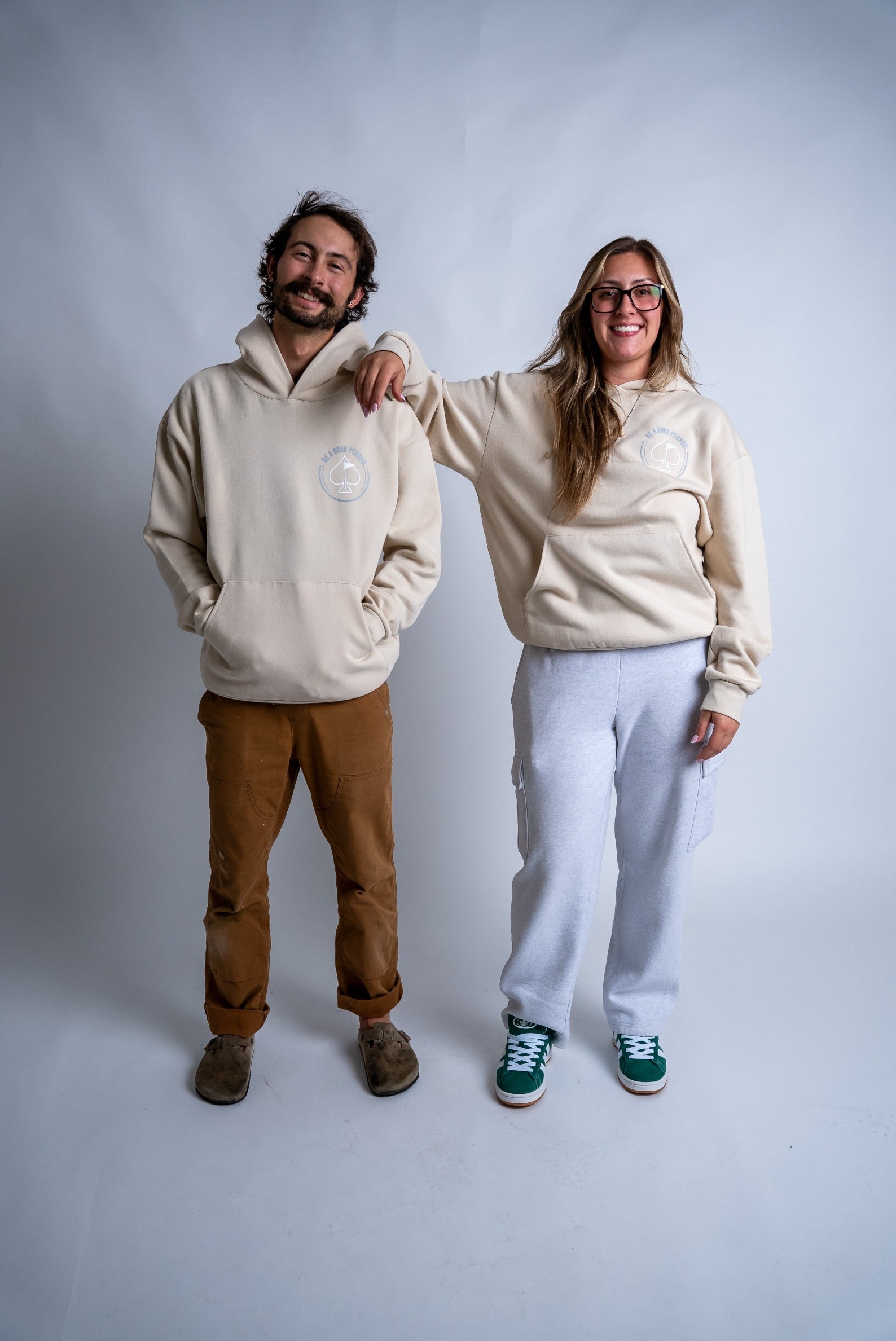 Cream hoodie featuring "Be A Good Person" design, worn by two smiling models in casual attire.