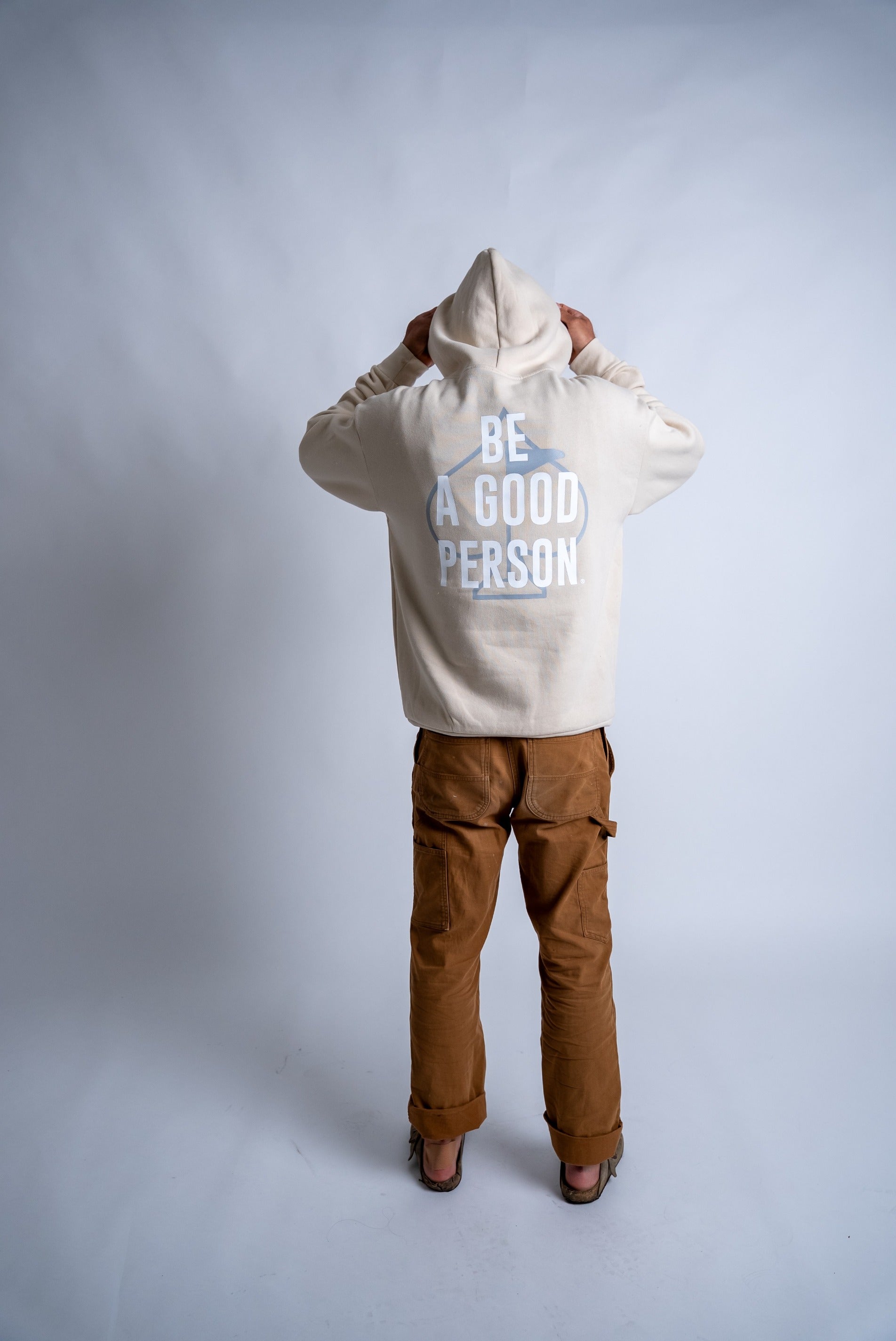 Cream hoodie with "Be A Good Person" printed on the back, worn by a person facing away, paired with brown pants.