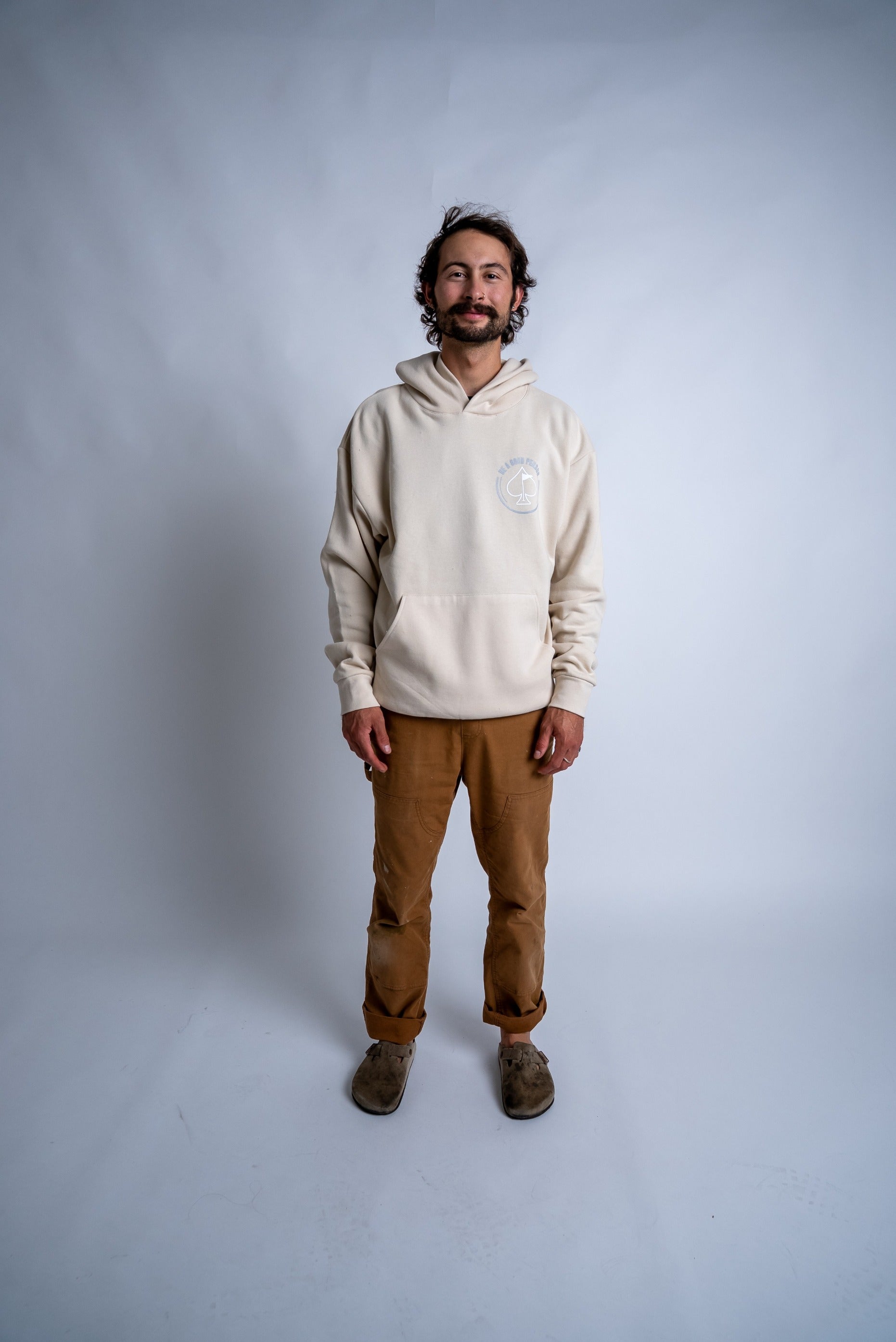 Cream hoodie featuring "Be A Good Person" design, worn by a smiling man standing against a neutral backdrop.