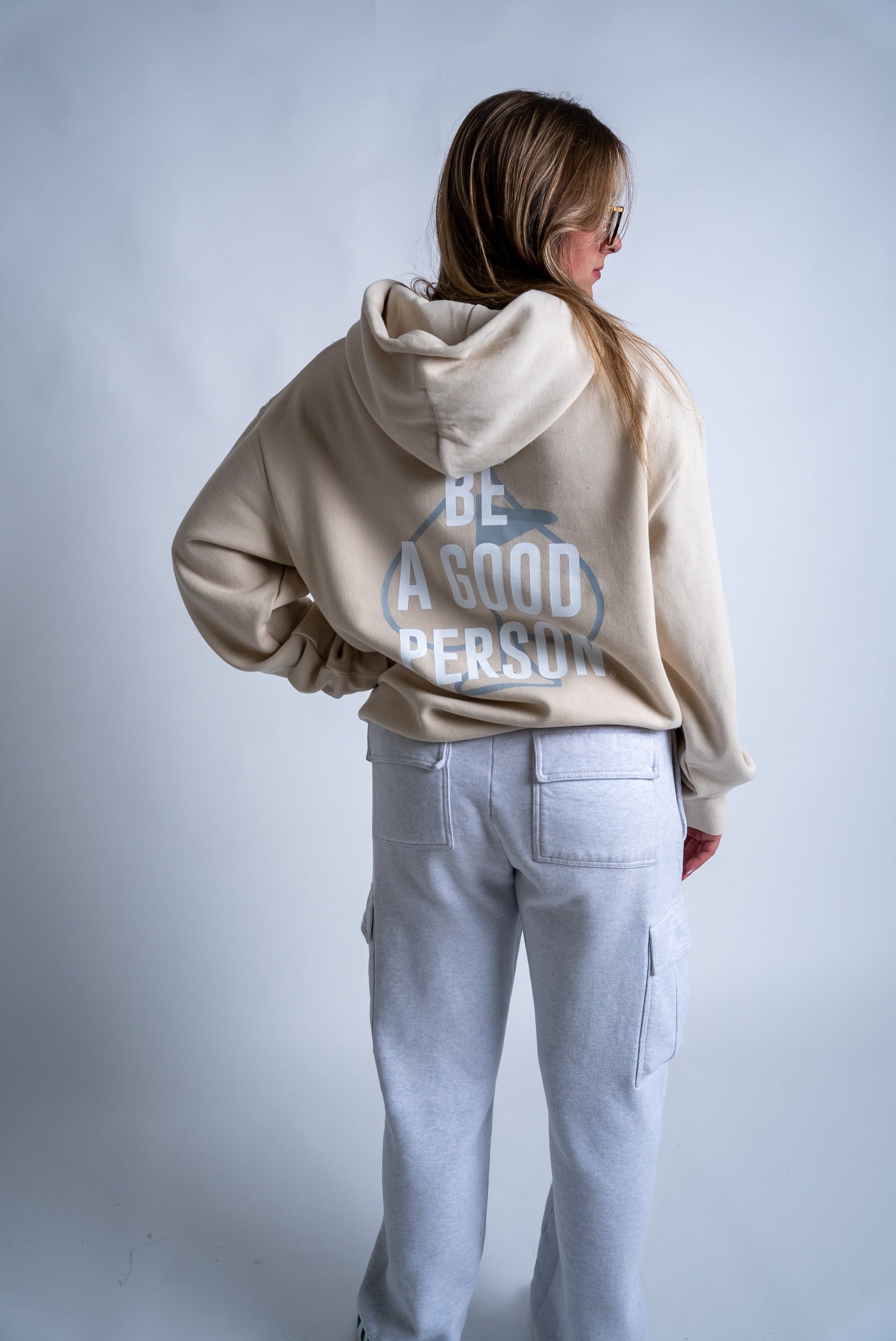 Cream hoodie with "Be A Good Person" text on the back, paired with gray cargo pants, showcasing a relaxed style.