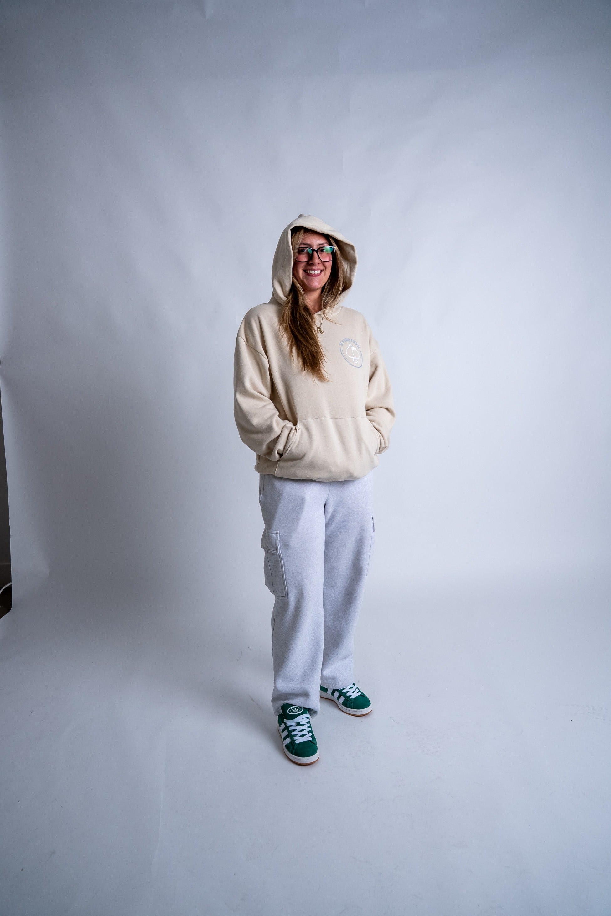 Cream hoodie featuring "Be A Good Person" design, paired with gray sweatpants and green sneakers, worn by a smiling woman.