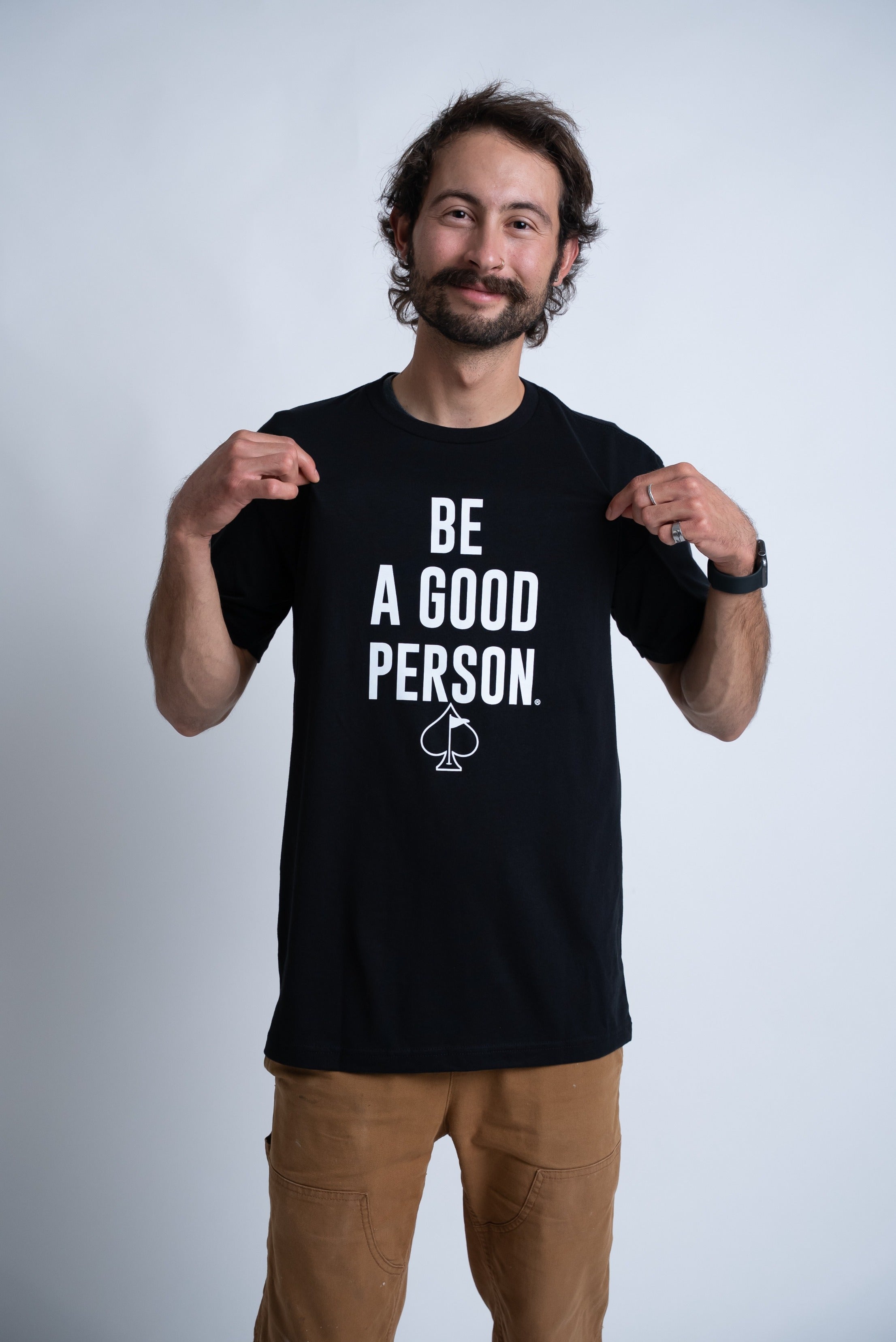 Man wearing a black Be A Good Person X Pins and Aces signature t-shirt, smiling and pointing at the shirt's message.