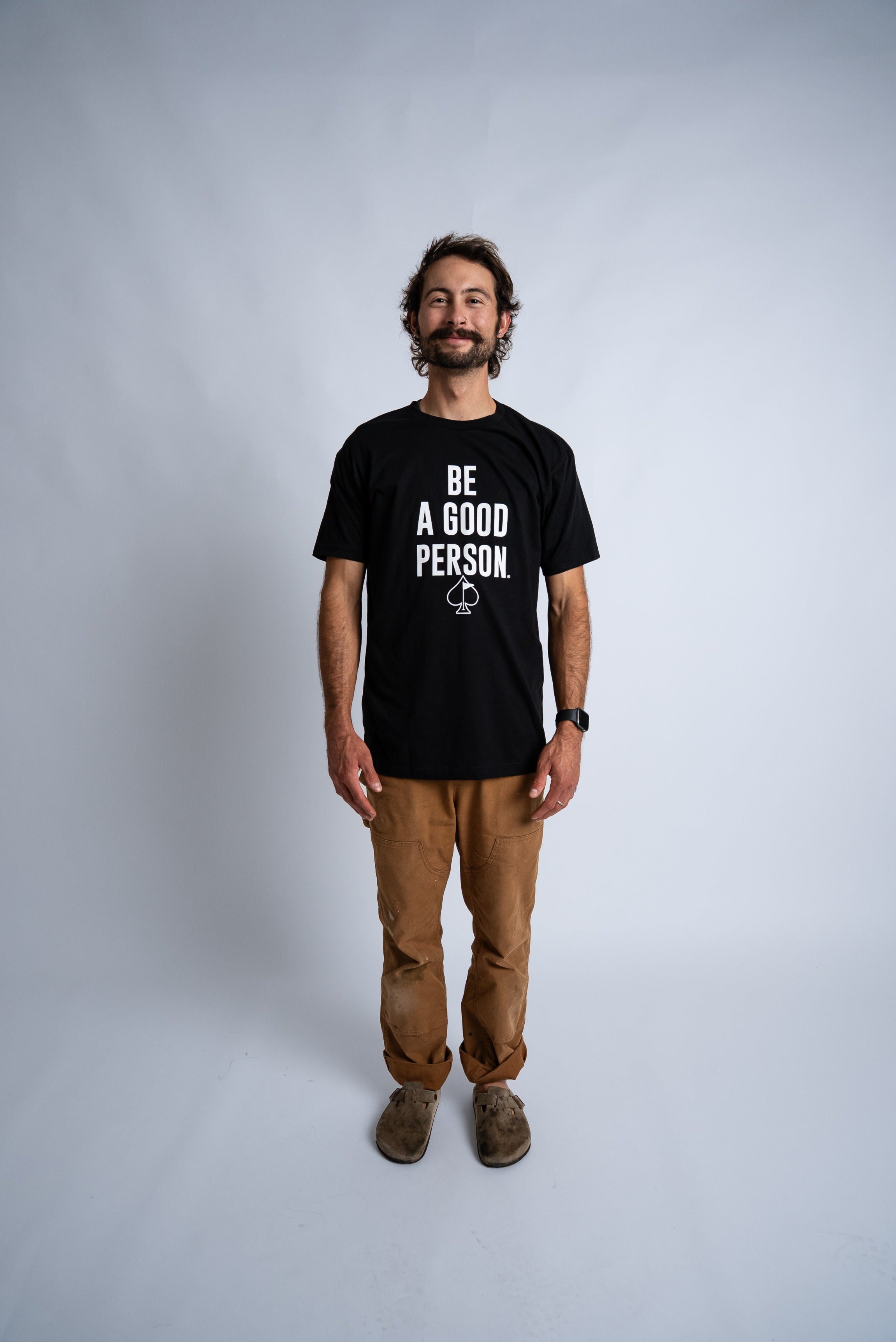 Black t-shirt featuring the phrase "Be A Good Person" in bold white letters, worn by a man standing confidently.