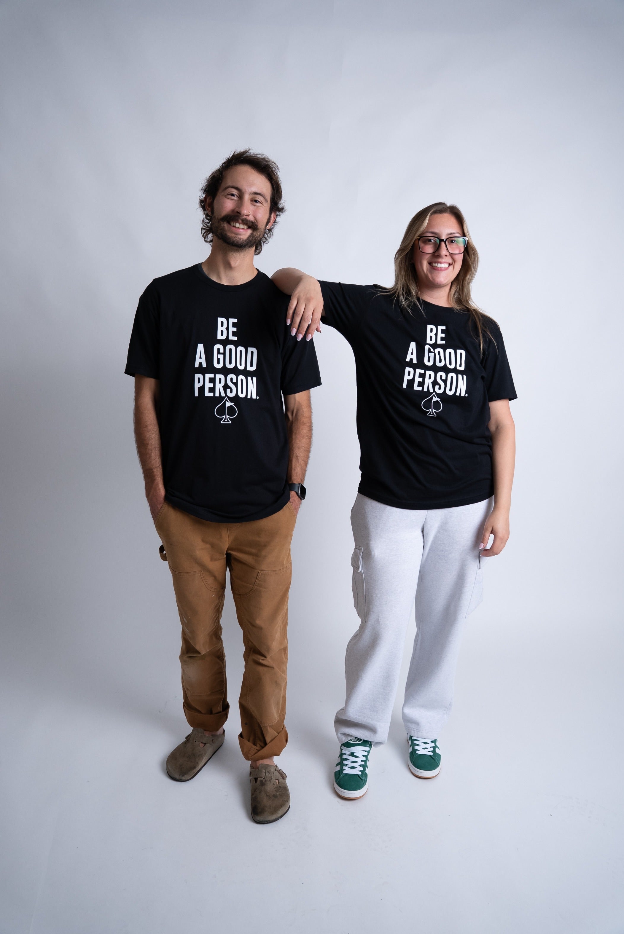 Black "Be A Good Person" t-shirts worn by a man and woman, showcasing a bold, positive message for golfers.