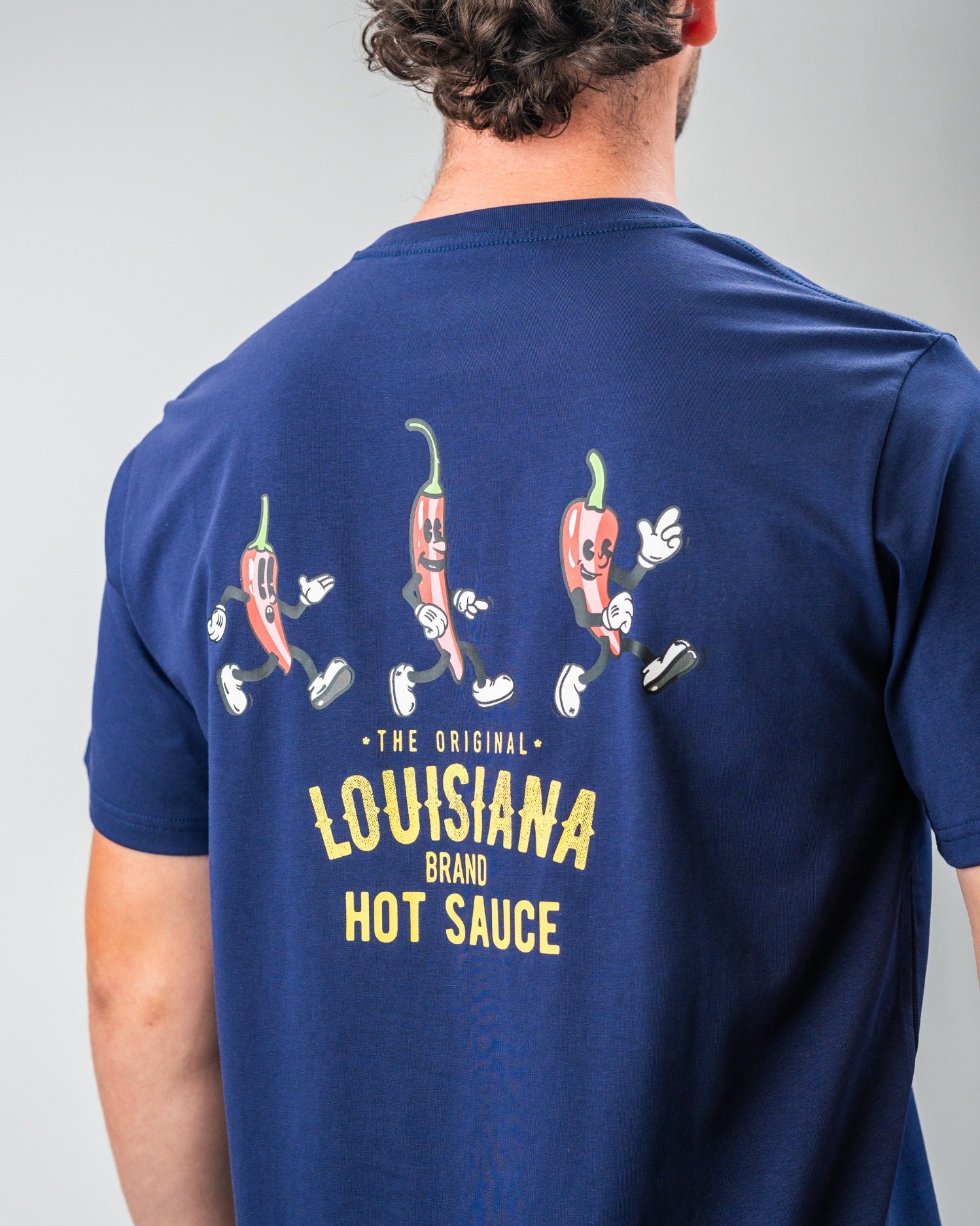 Navy tee featuring playful dancing pepper graphics and "Louisiana Hot Sauce" branding on the back. Perfect for hot sauce fans.