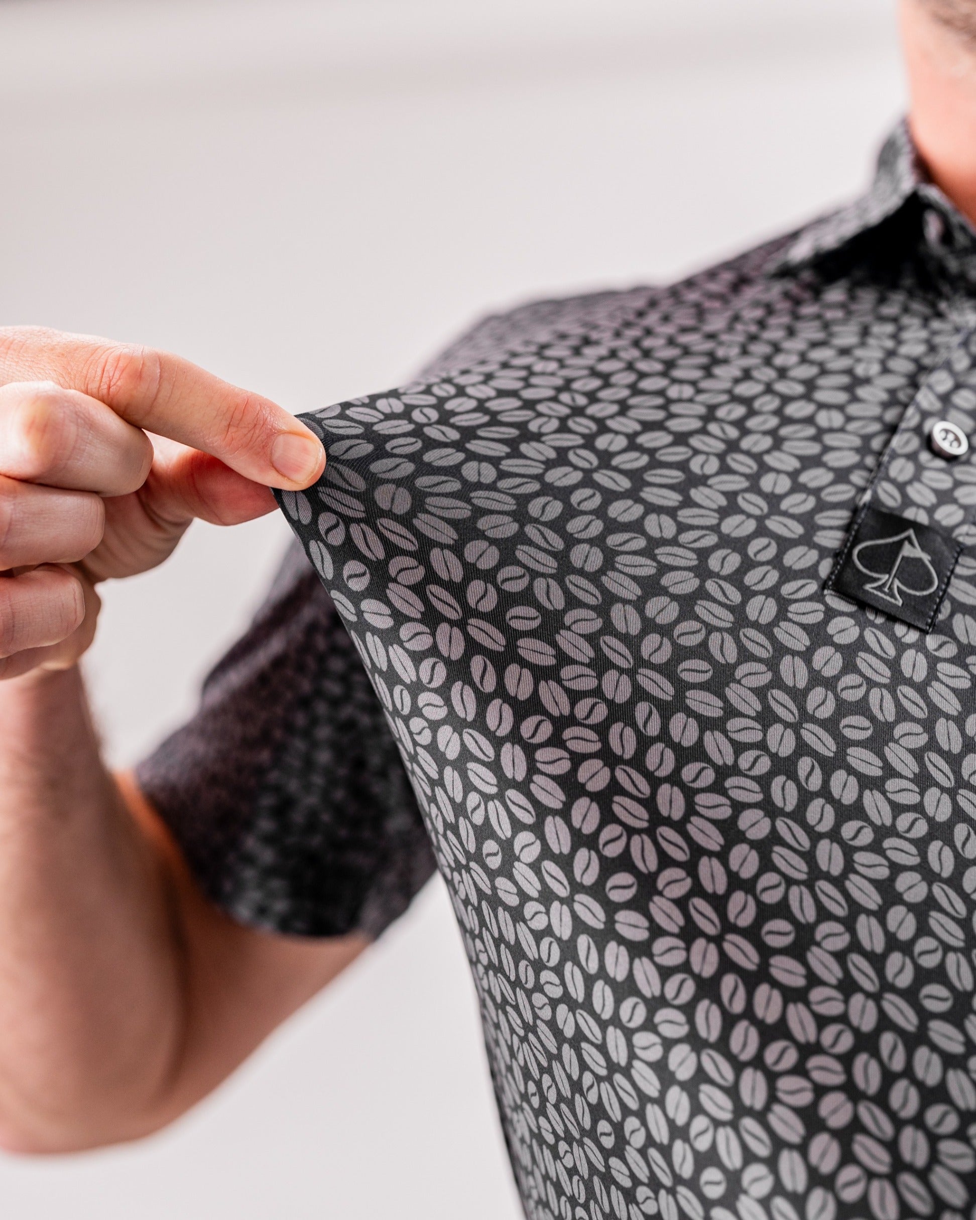 Close-up of a person holding the sleeve of a black polo shirt featuring a coffee bean pattern, showcasing its unique design.