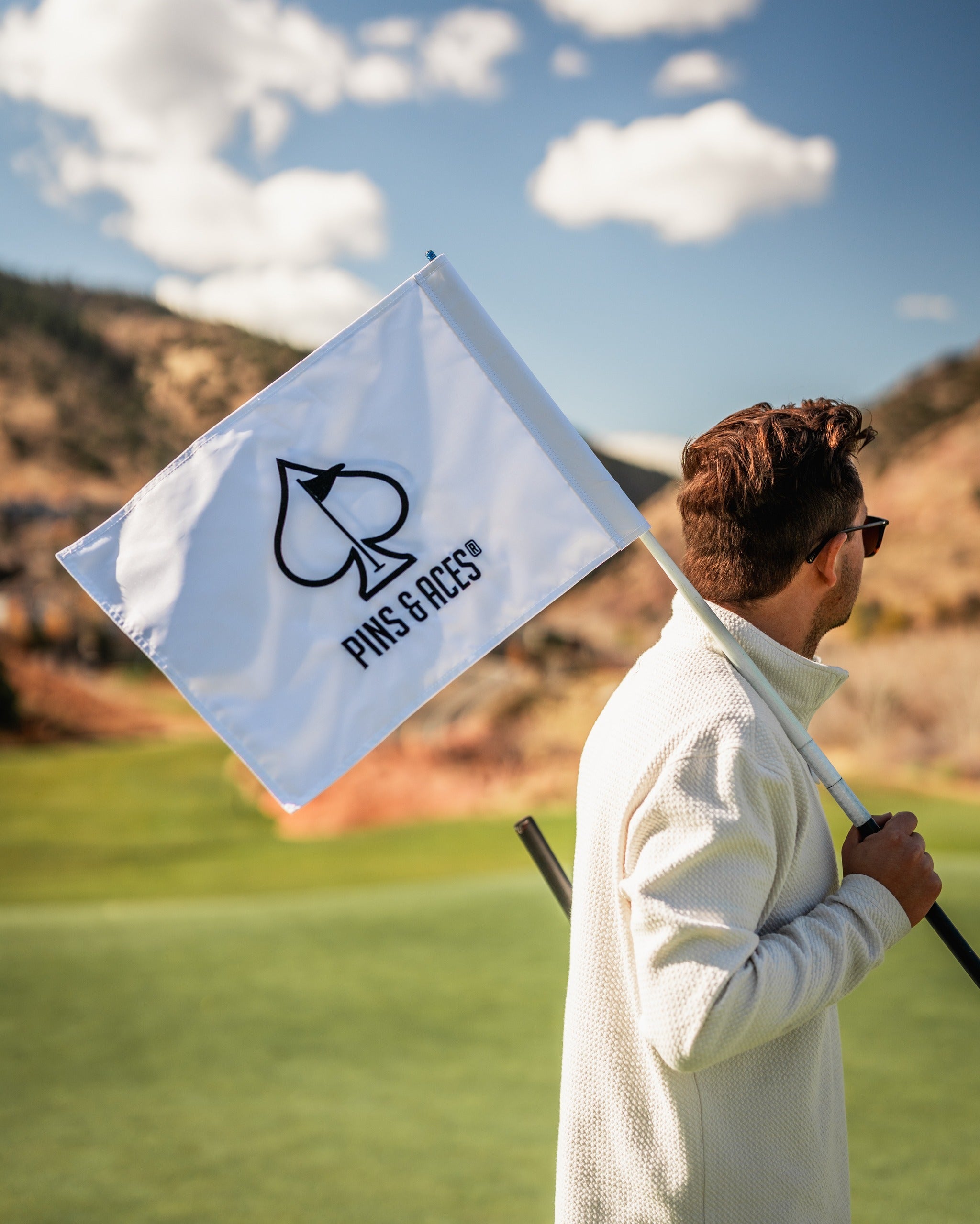 A golfer holds a white Pins and Aces flag featuring a spade logo, set against a scenic golf course backdrop.