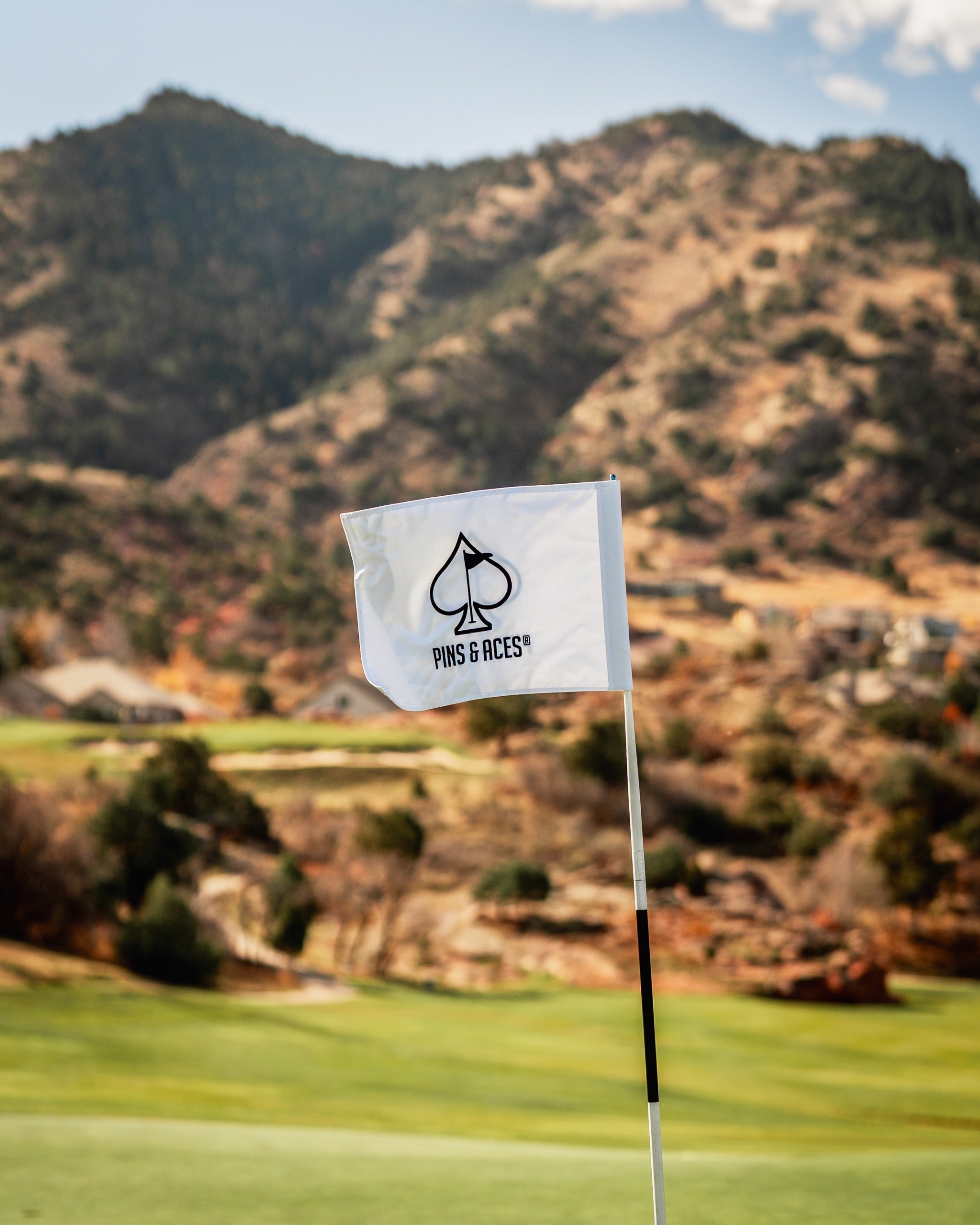 White golf flag featuring the Pins and Aces logo, set against a scenic mountainous landscape.