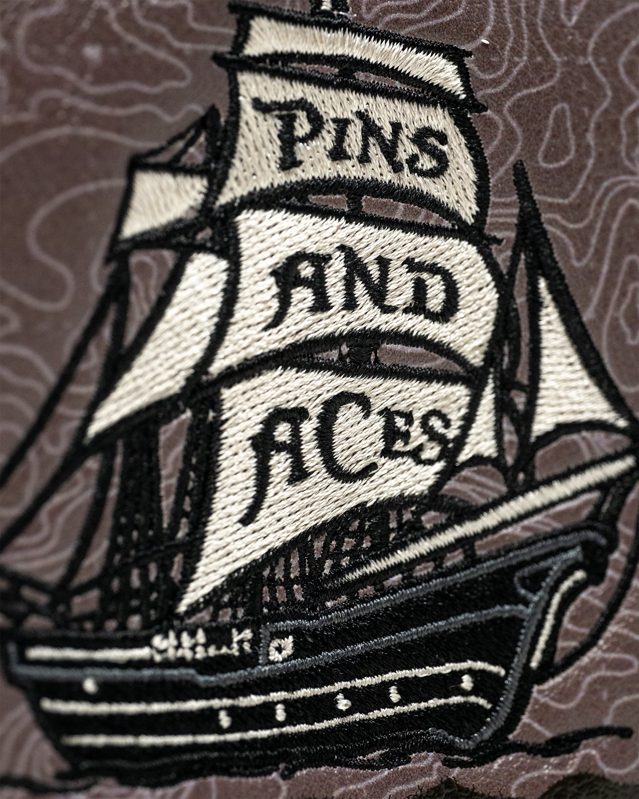 Bold embroidered patch of a pirate ship with "PINS AND ACES" banners, black and white colors, on textured background.