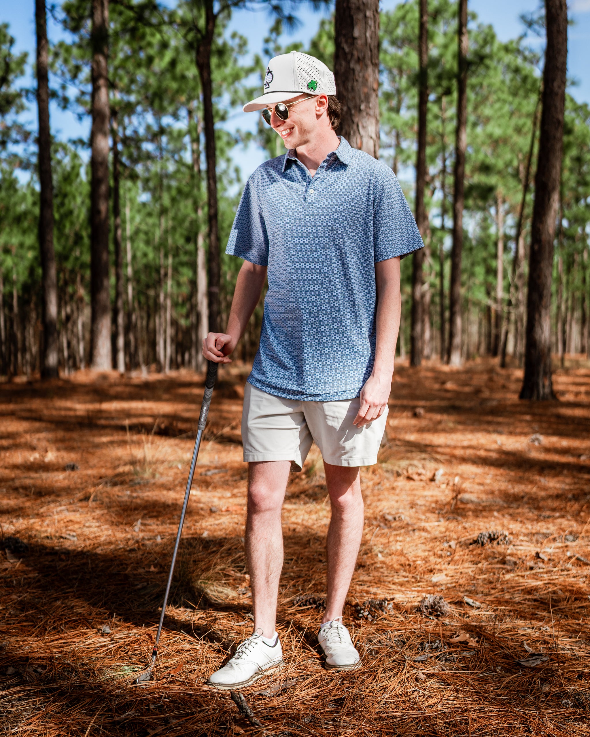 A golfer stands in a pine forest wearing a blue patterned polo shirt and beige shorts, holding a golf club.