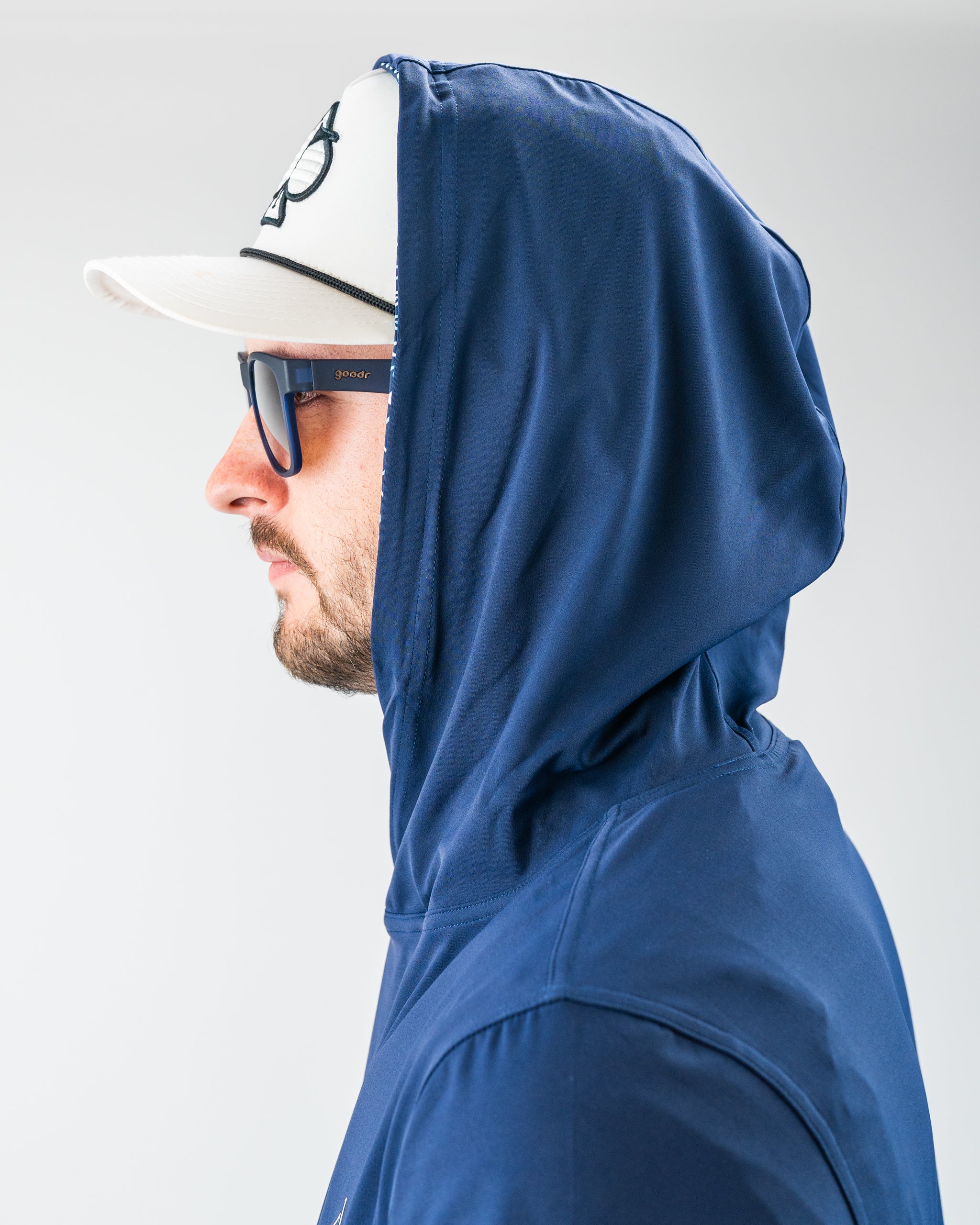 Man wearing a navy blue hooded jacket, white cap, and blue sunglasses, showcasing bold, stylish golf apparel.