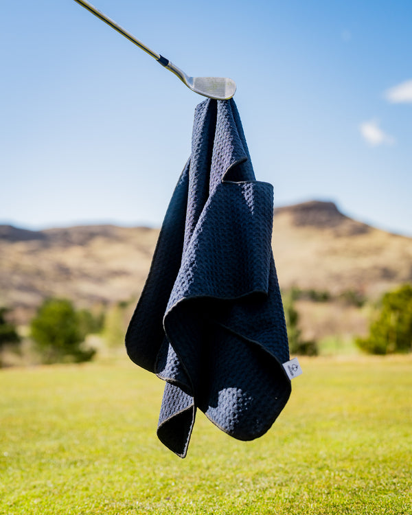 Black textured golf towel hanging from a golf club, with a scenic outdoor background and mountains.