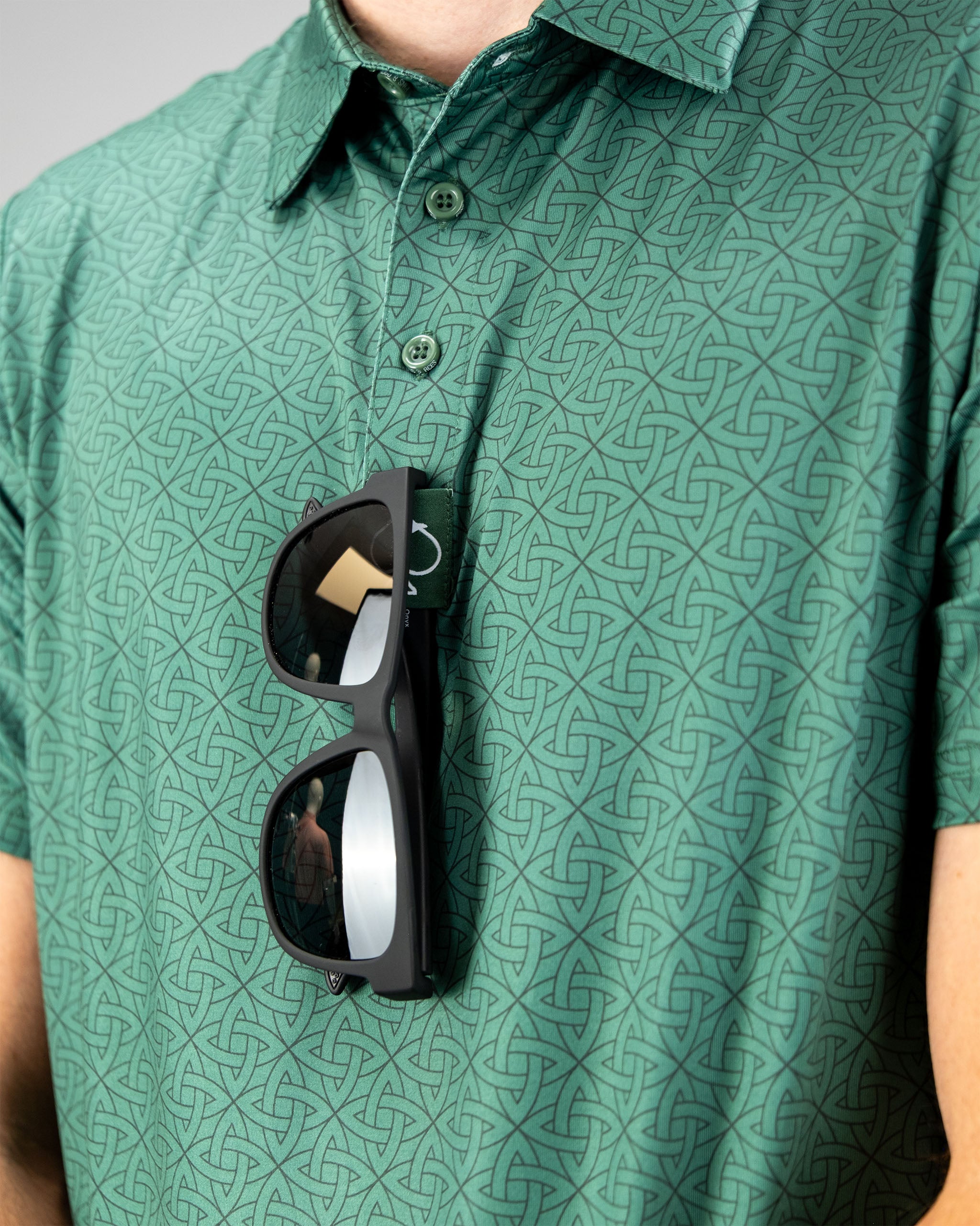 Model wearing an emerald green polo shirt with a geometric pattern, sunglasses resting on the collar.