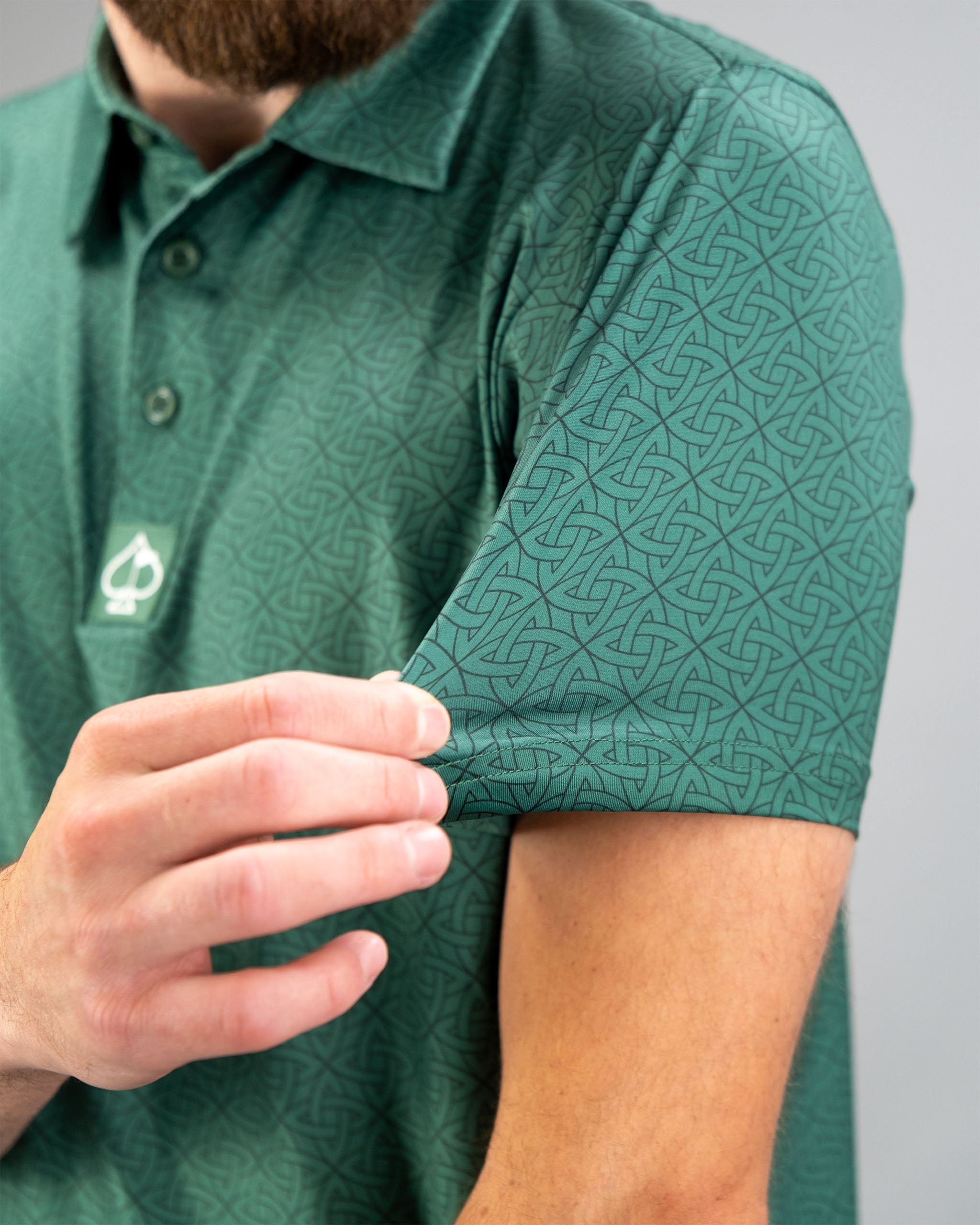 Close-up of a man adjusting the sleeve of a green polo shirt featuring an intricate geometric pattern.