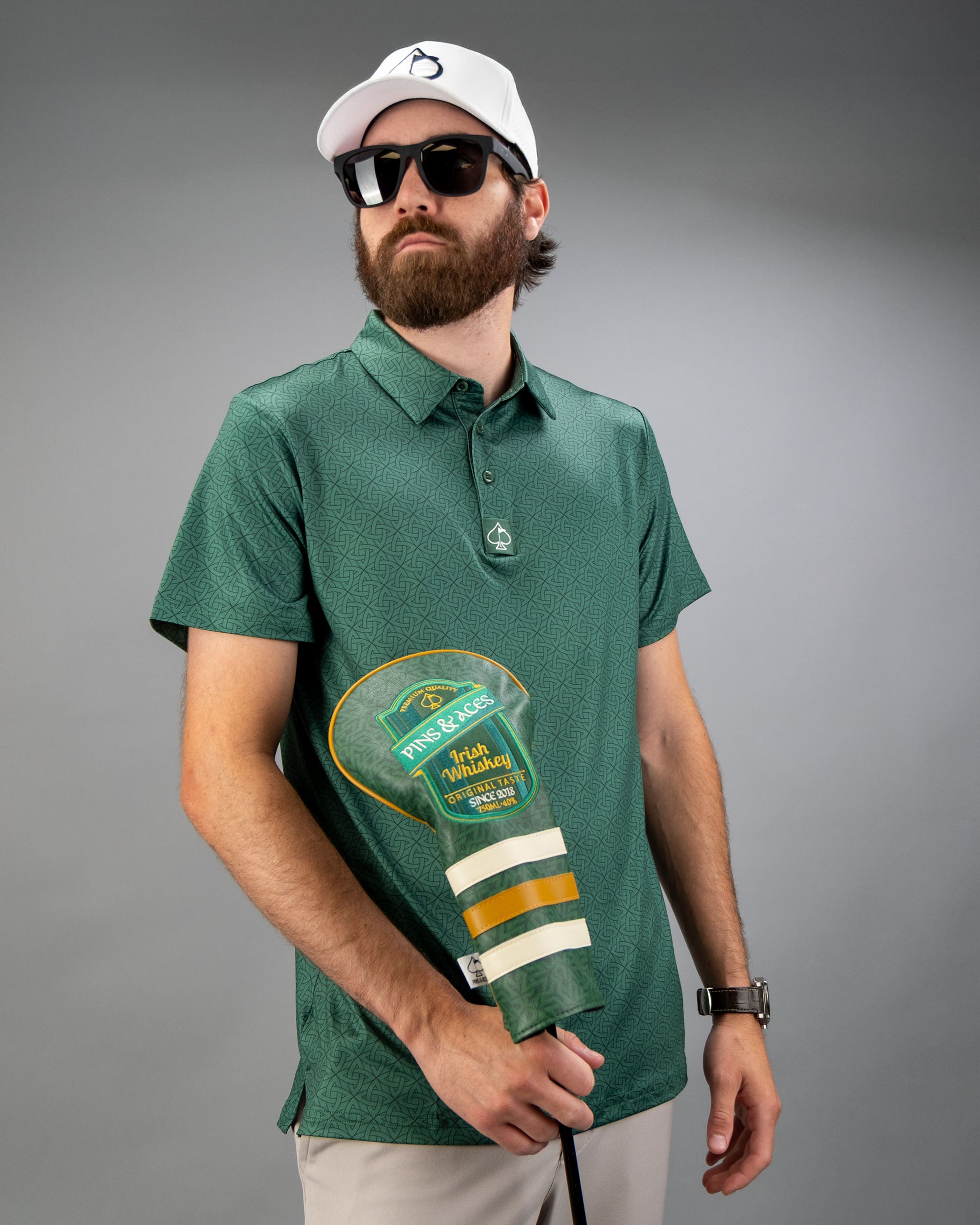 Man wearing an emerald green polo shirt with a geometric pattern, holding a golf club cover, and wearing sunglasses and a cap.