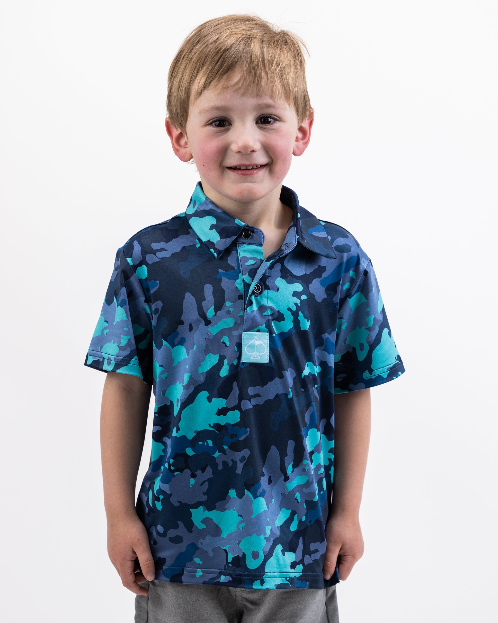 Young boy wearing a blue electric camo pattern polo shirt with short sleeves and a collar, smiling against a plain white background.