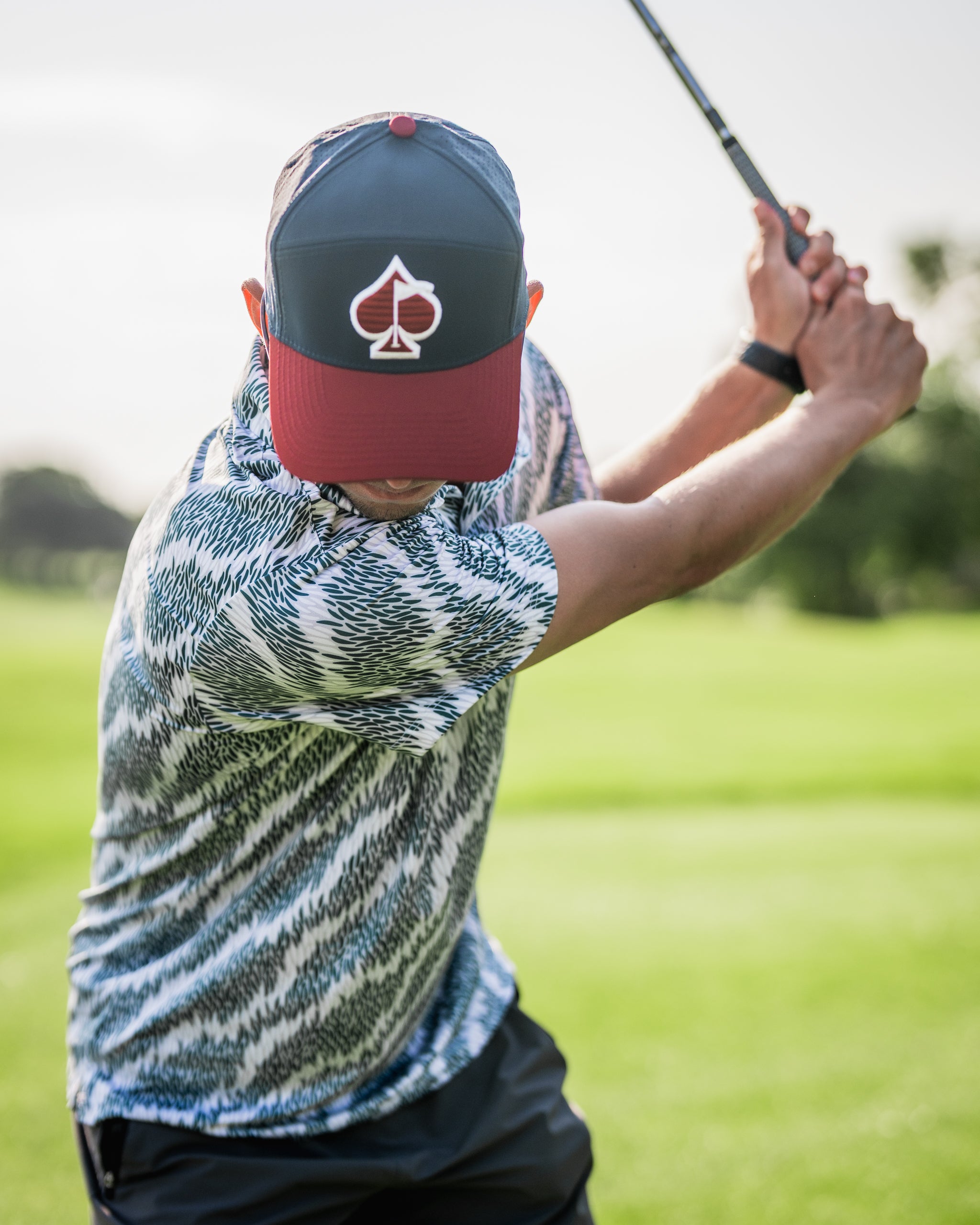 A golfer swings a club wearing a patterned shirt and a cap featuring a spade logo, set against a lush green background.