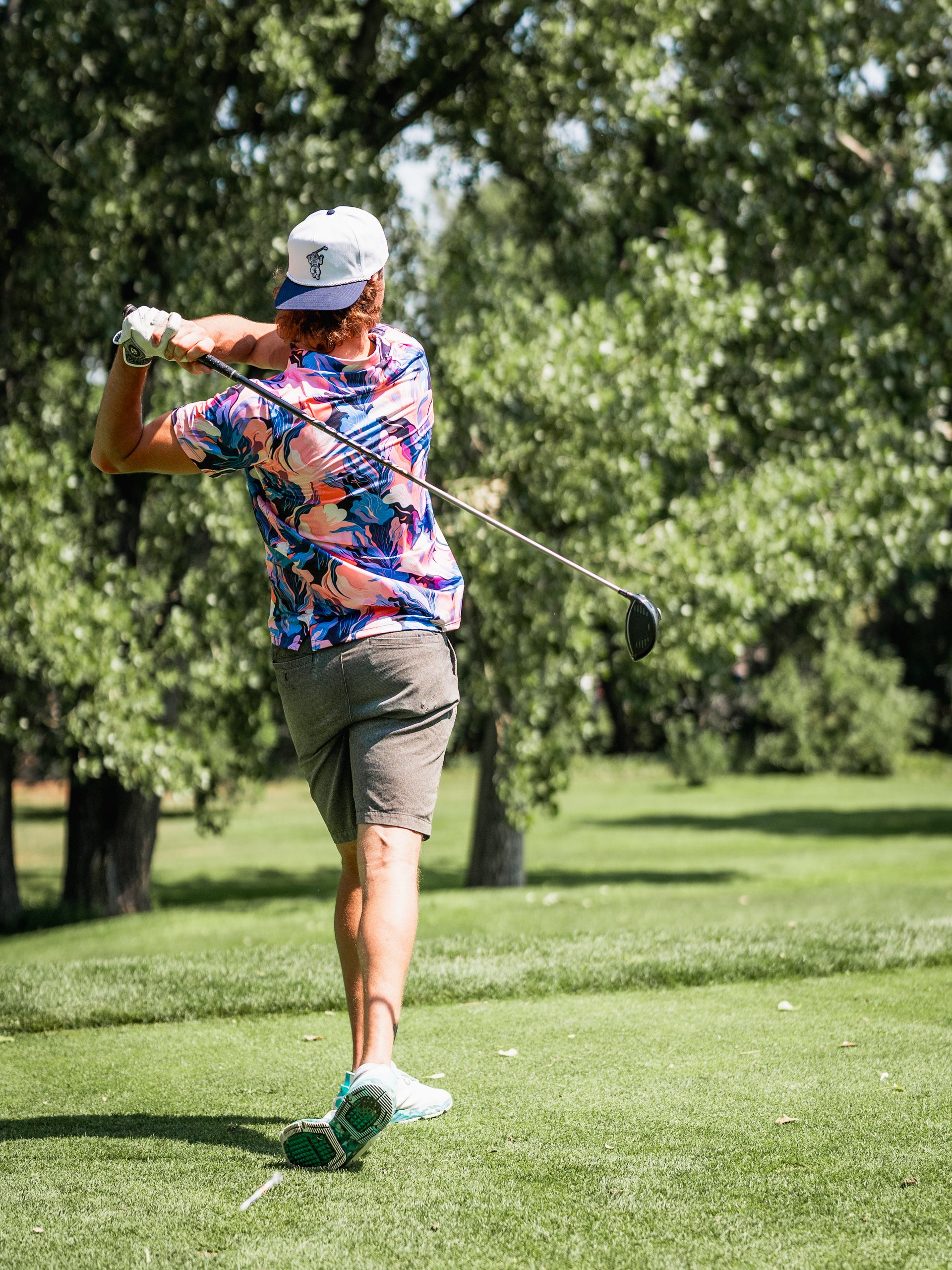 A golfer swings a club on the course, wearing a vibrant melting floral shirt and shorts, surrounded by trees.