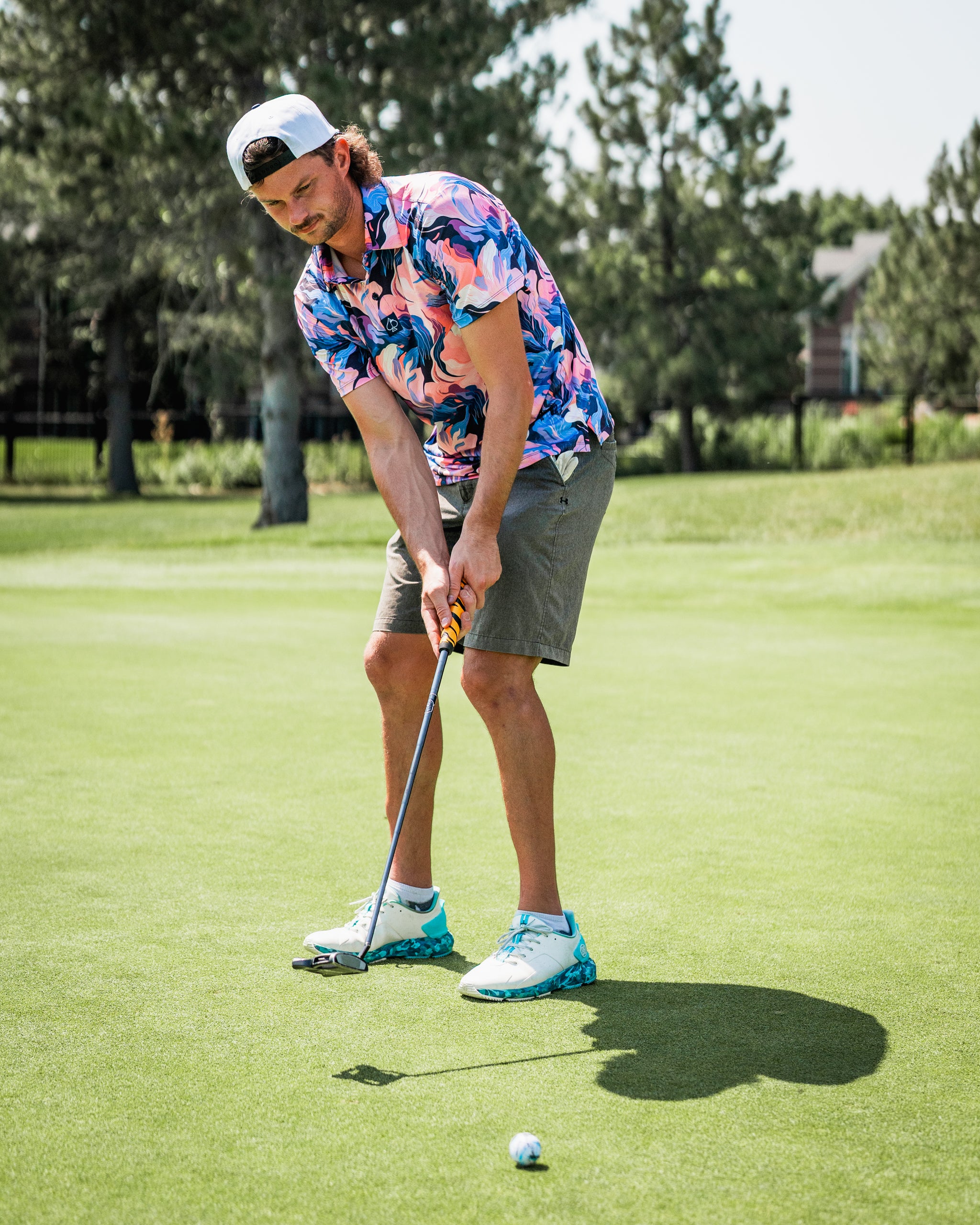 A golfer in a vibrant melting floral shirt and shorts prepares to putt on a sunny green course.