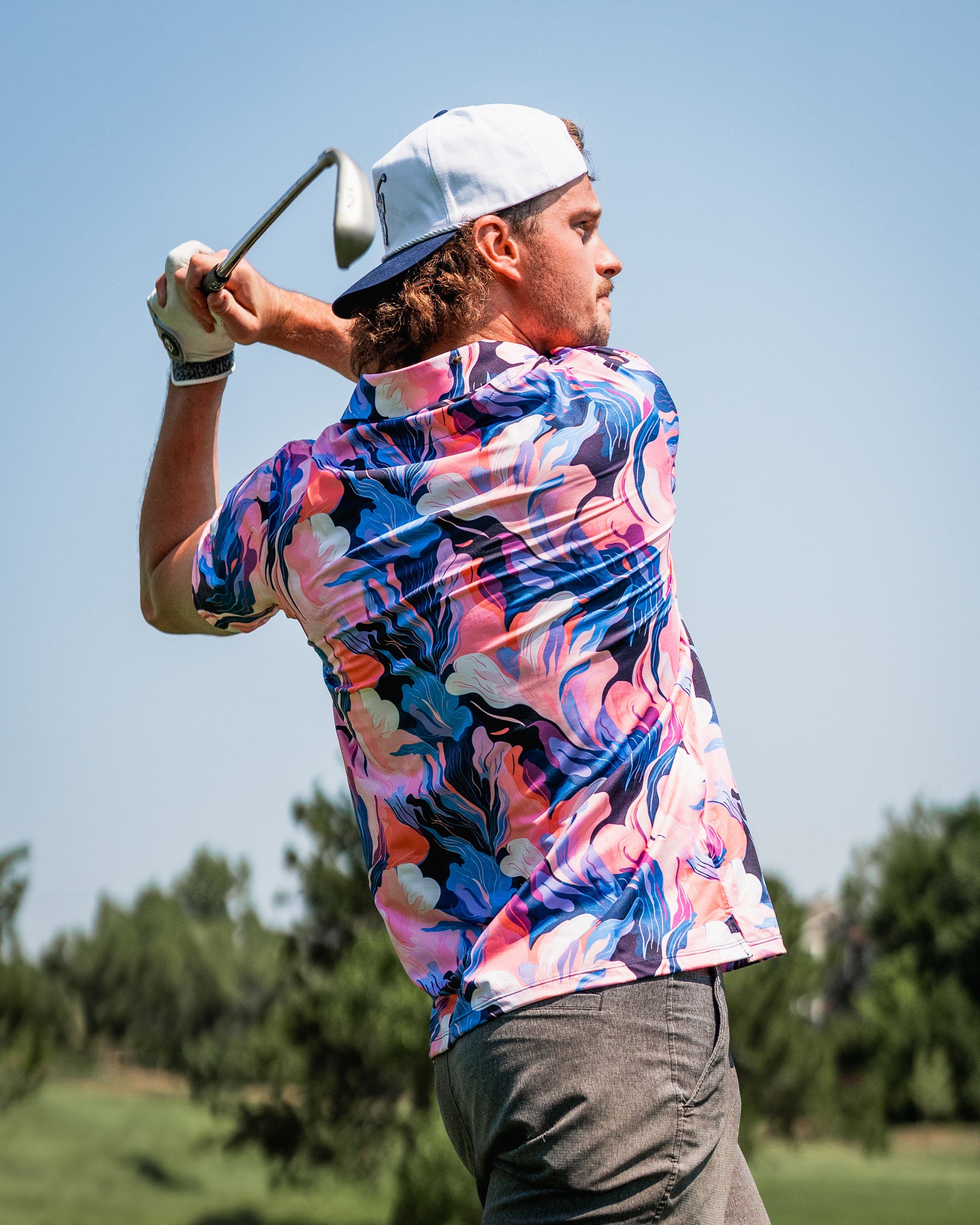 A golfer swings a club, wearing a vibrant melting floral patterned shirt against a clear blue sky.
