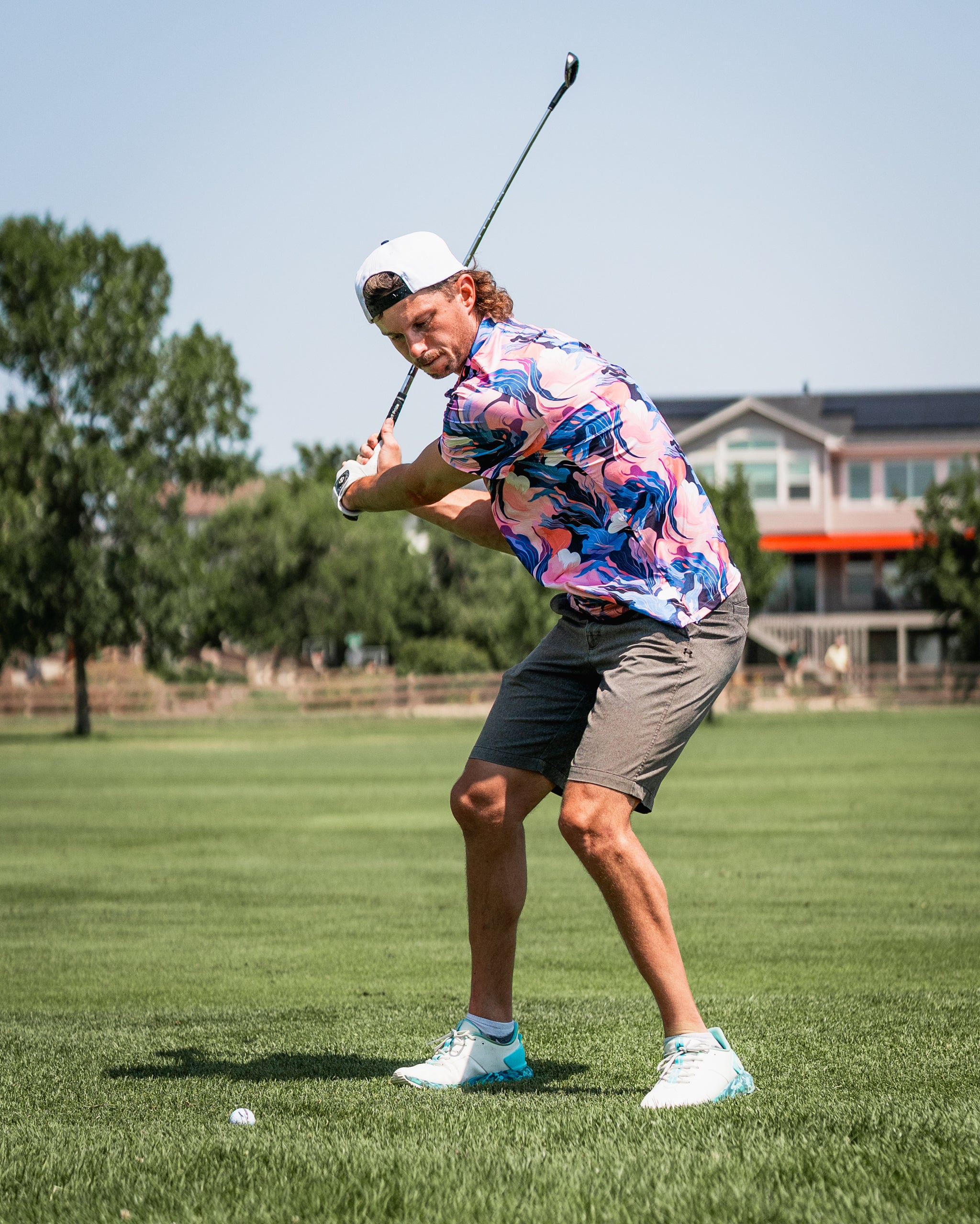 A golfer swings a club on the course, wearing a vibrant melting floral shirt and gray shorts, showcasing bold style.