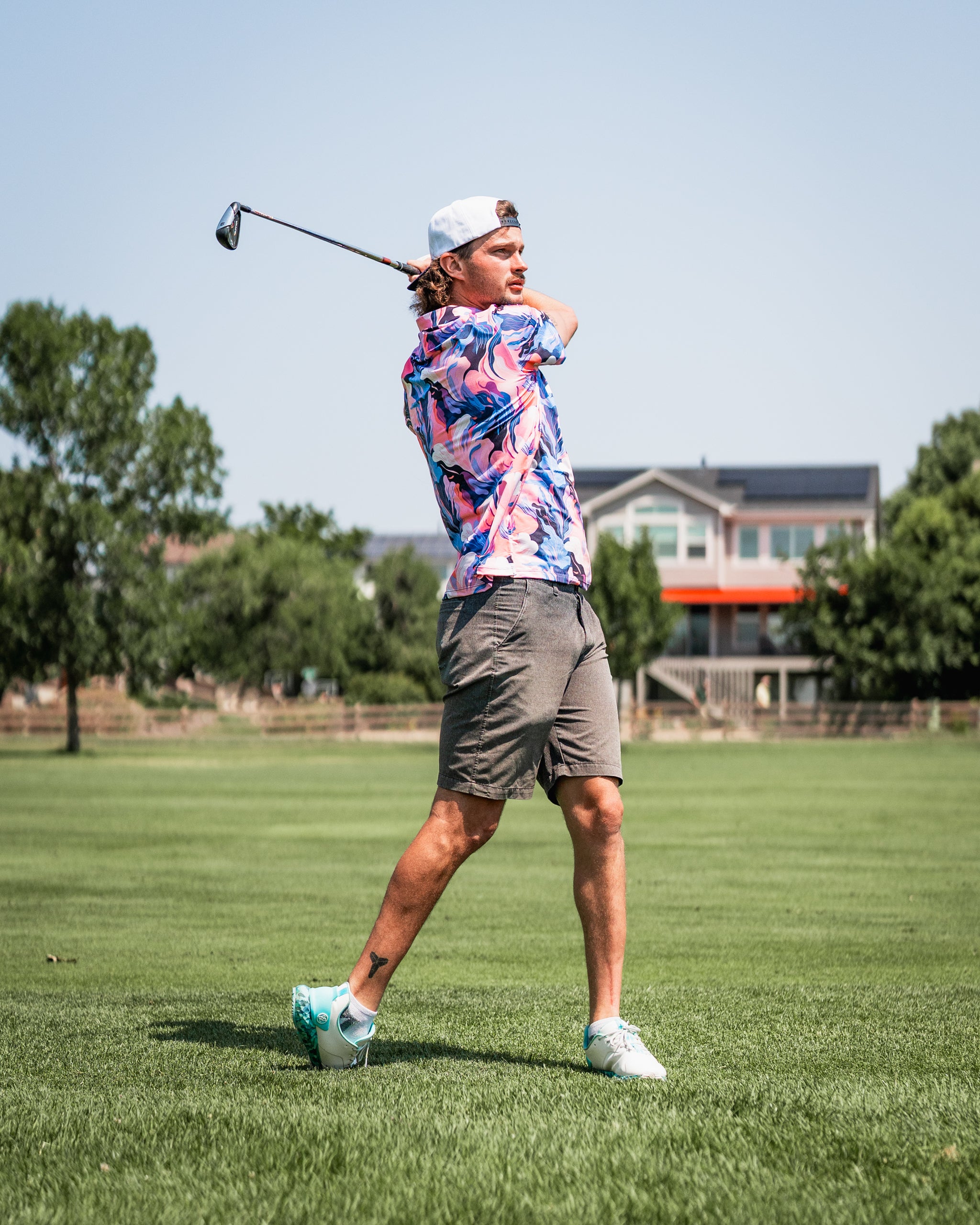 A golfer swings a club on a sunny day, wearing a vibrant melting floral shirt and shorts on a green course.