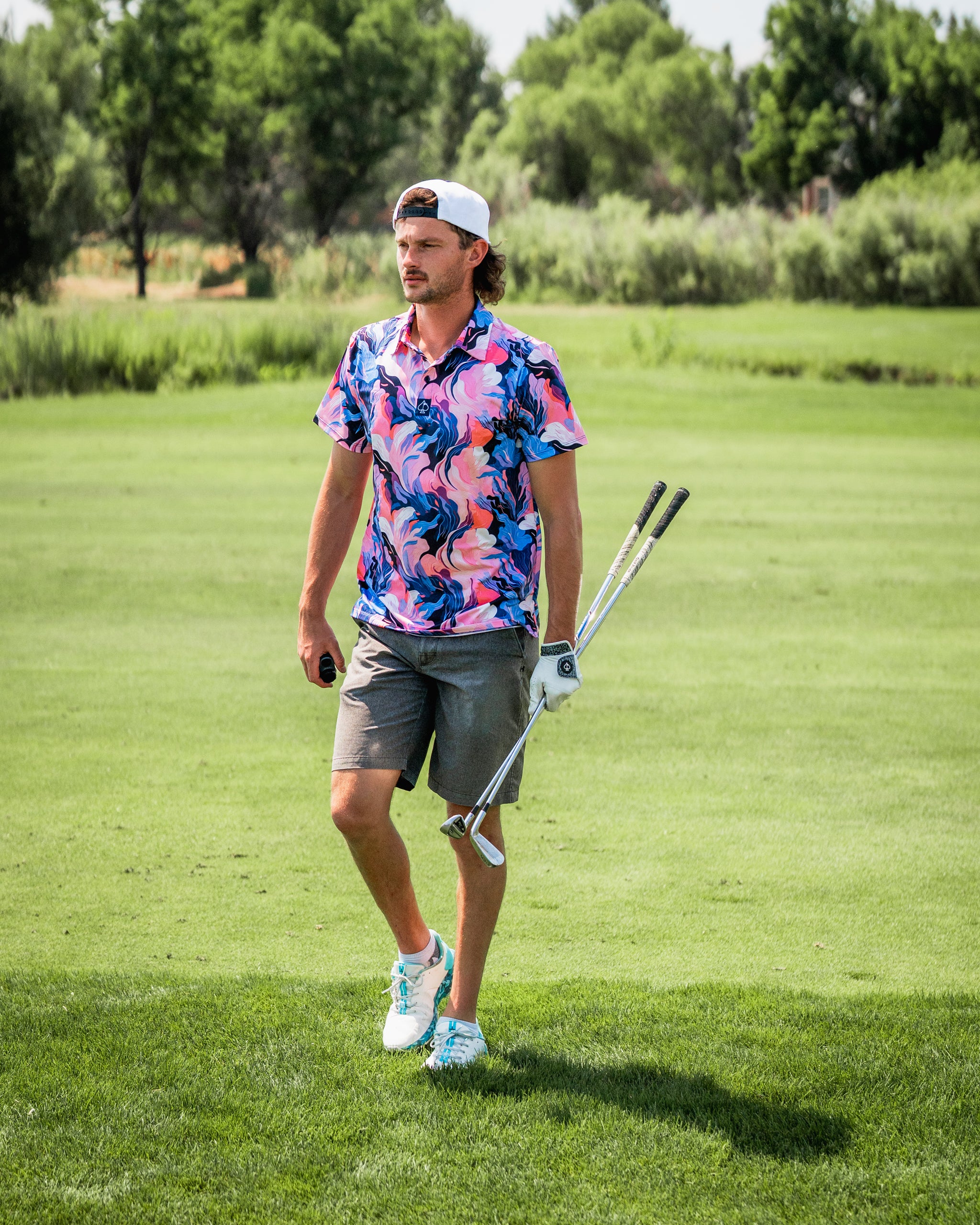 A golfer walks on the course wearing a vibrant melting floral shirt and gray shorts, holding golf clubs.